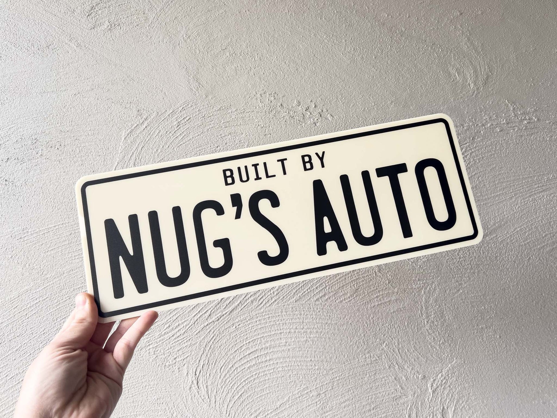 Hand holding a beige acrylic plate with 'Built by Nug's Auto' in black text