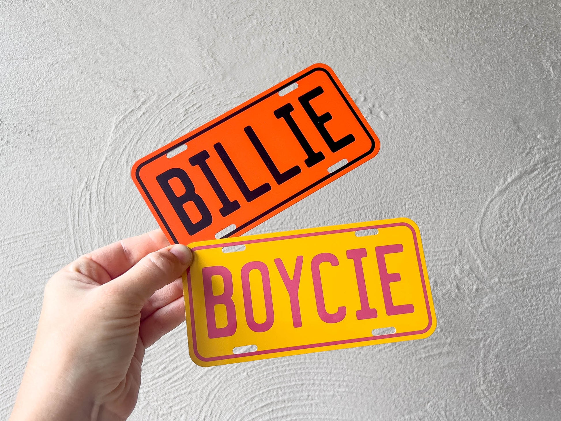 Two mini license plate-style signs with 'BILLIE' and 'BOYCIE' text held by a hand against a light grey background.