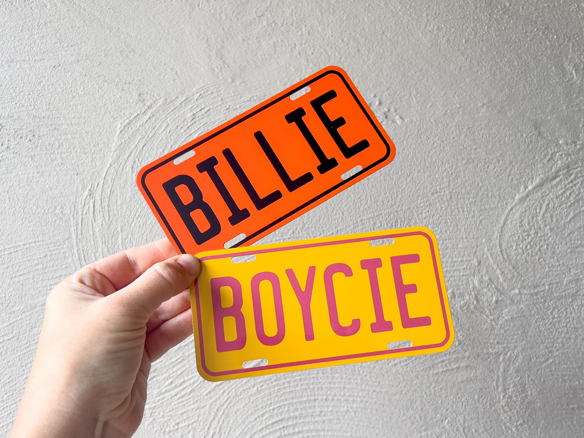 Two mini license plate-style signs with 'BILLIE' and 'BOYCIE' text held by a hand against a light grey background.