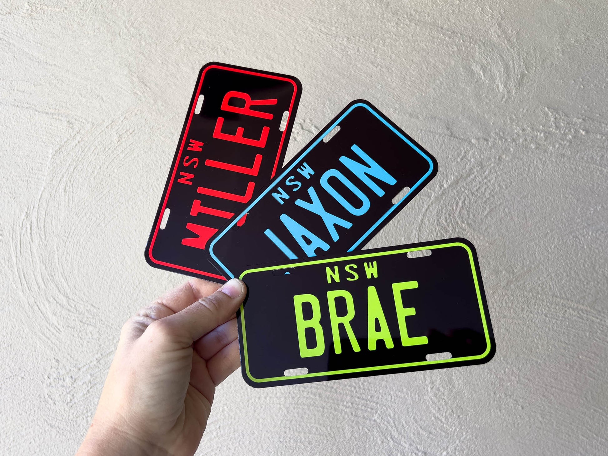 Three novelty mini number plates with text held by a hand against a light grey background