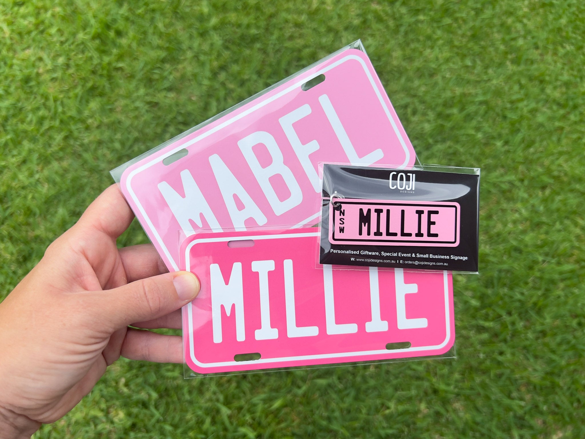 Pink license plates with 'MABEL' and 'MILLIE' text held by a hand on grass.