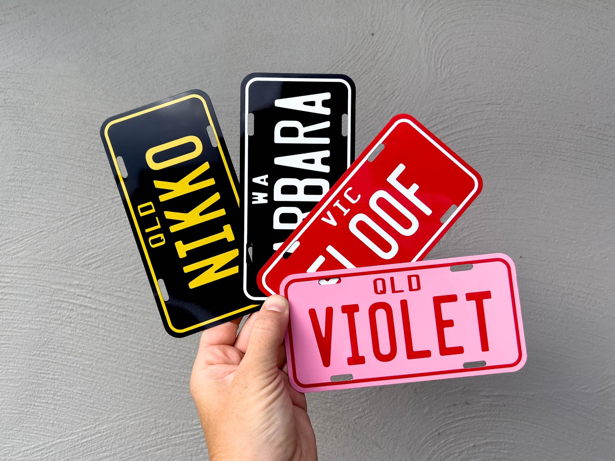 Four license plate-style name signs held by a hand against a grey background