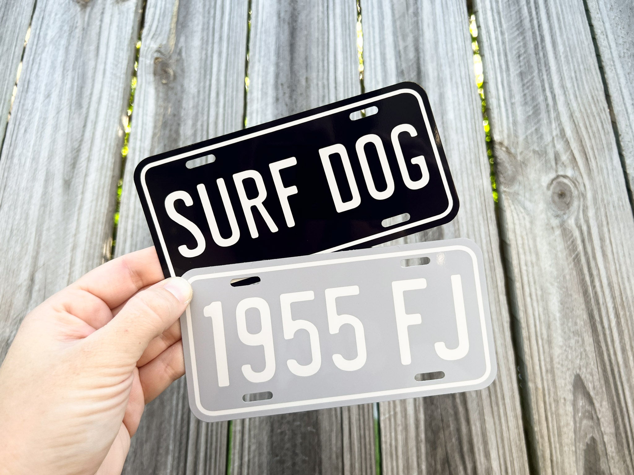 Two mini number plates, one black with 'SURF DOG' text and the other white with '1955 FJ' text, held against a wooden background.