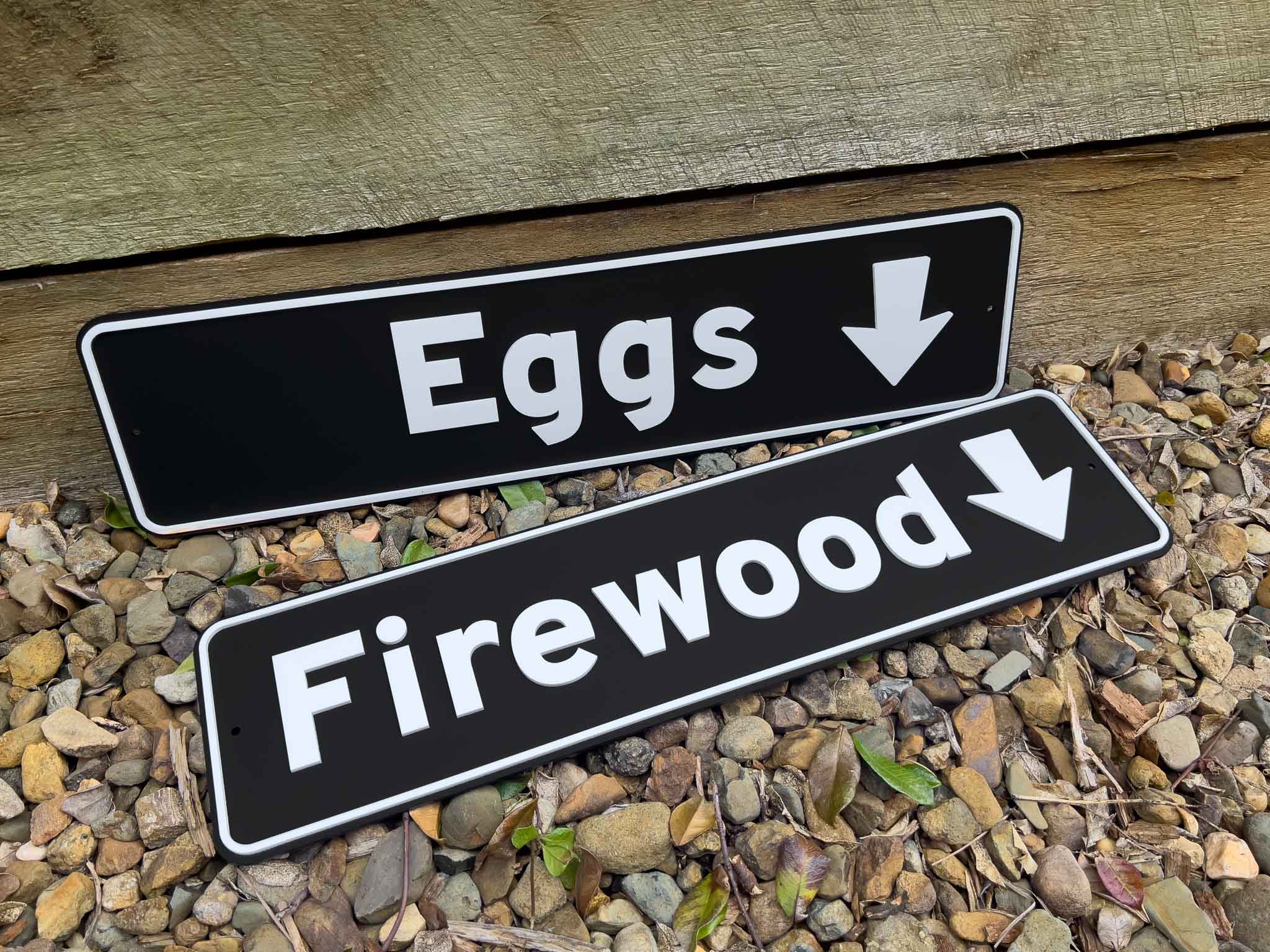 Two black and white highway style signs with the words 'Eggs' & 'Firewood' with a downward pointing arrow.