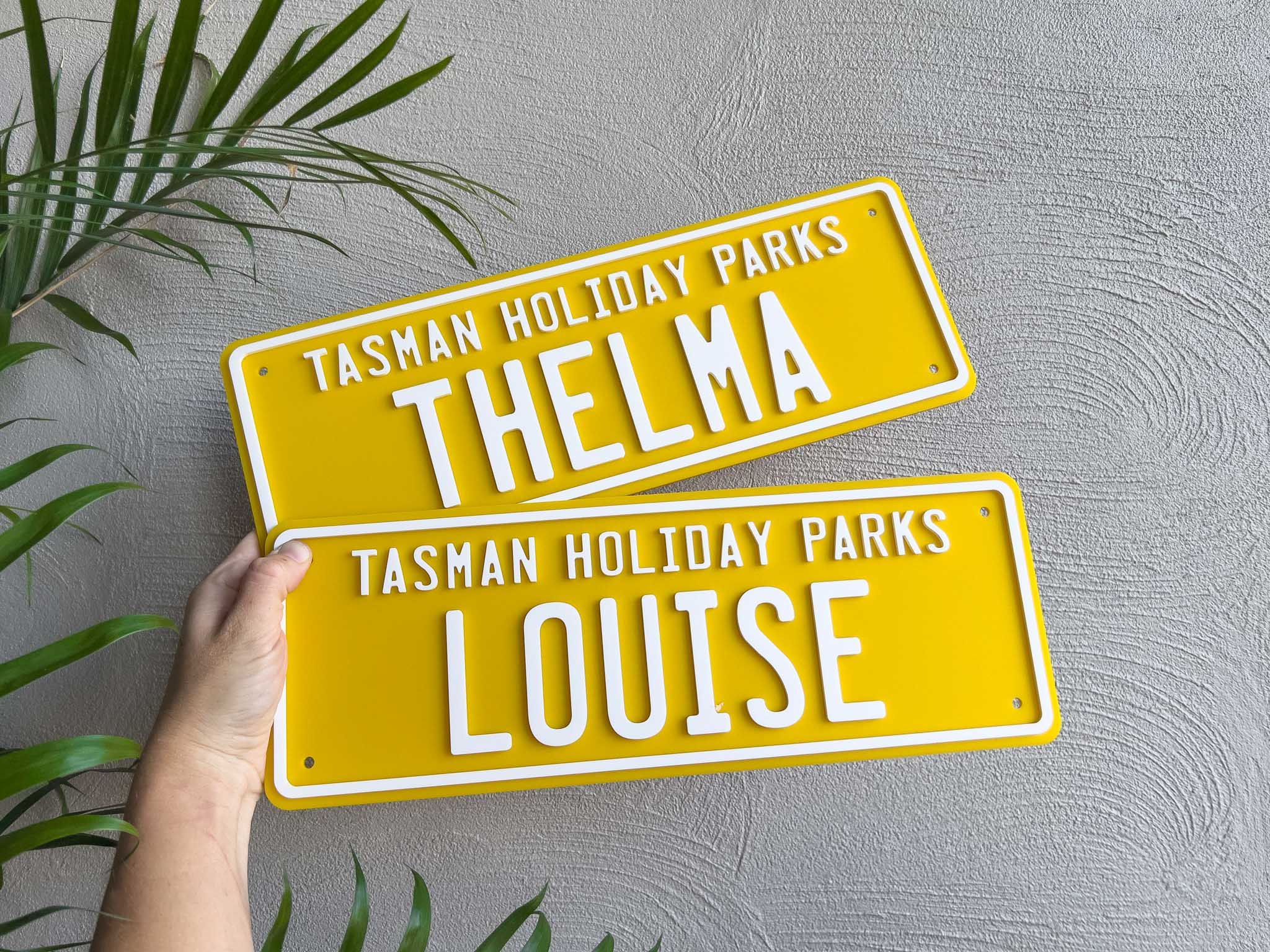 Two custom acrylic yellow and white signs with 'Tasman Holiday Parks' and names 'Thelma' and 'Louise' on a grey background.