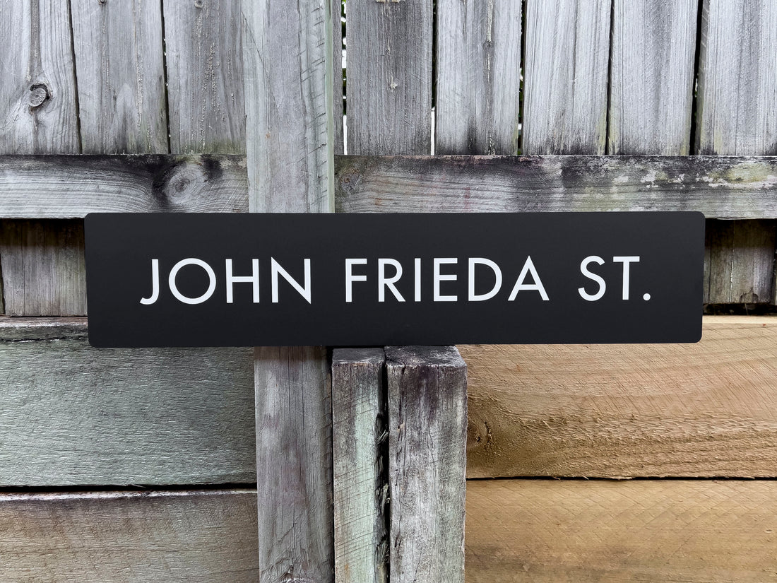 Custom street sign with 'JOHN FRIEDA ST.' text against a wooden background
