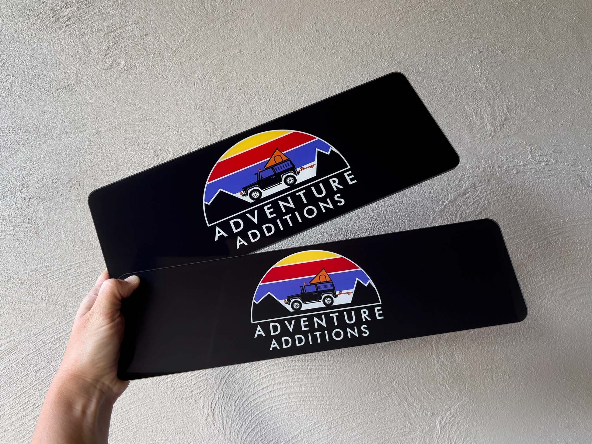 Two black glossy logo plates with the brand 'Adventure Additions' in standard and slim sizes.