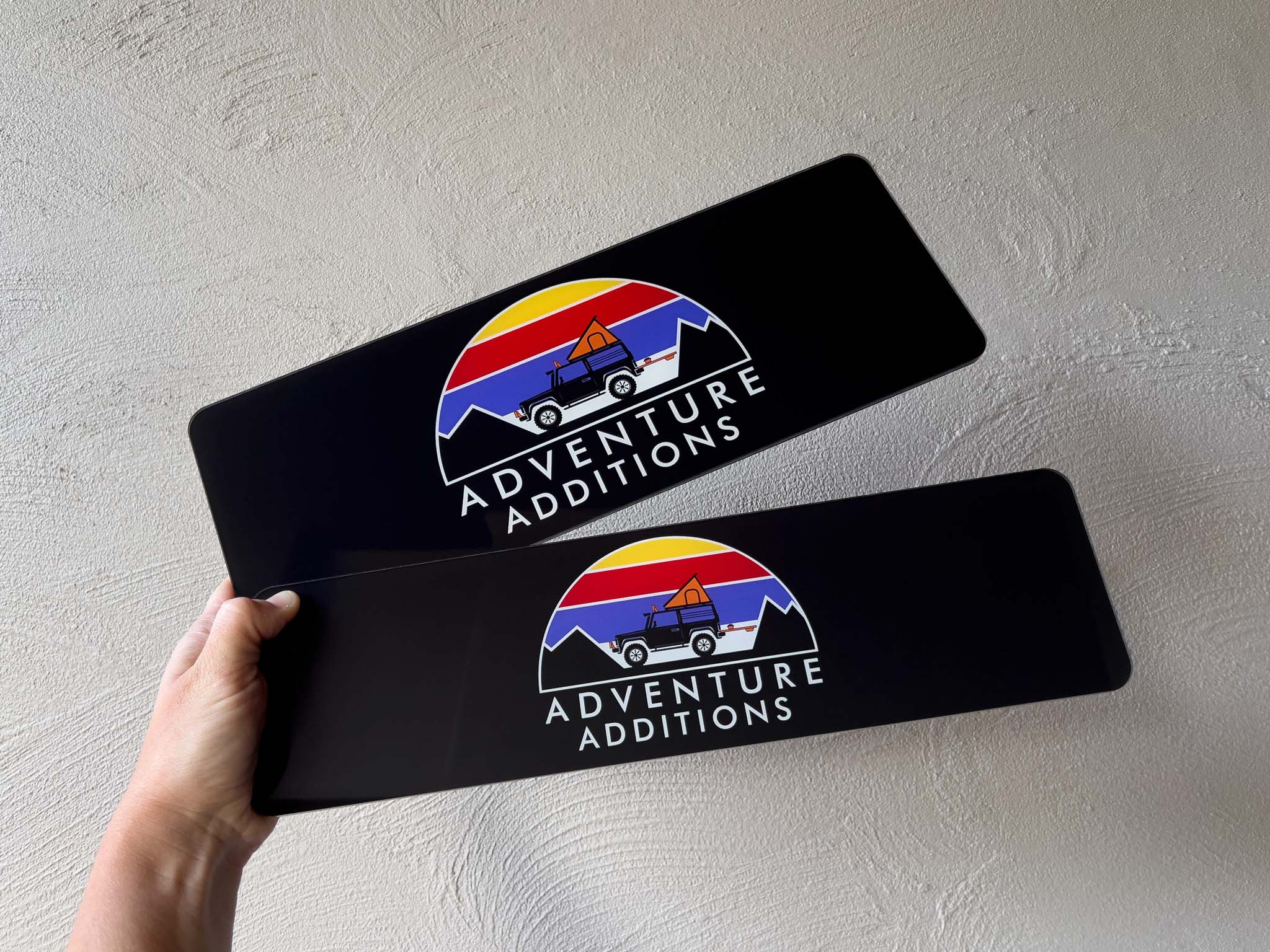 Two black glossy logo plates with the brand 'Adventure Additions' in standard and slim sizes.