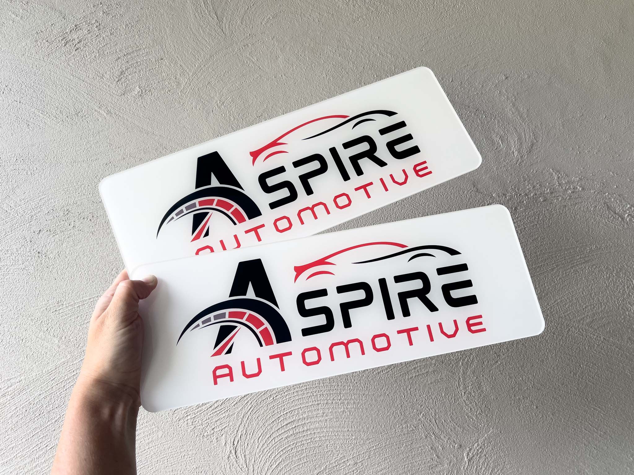 Two 'Aspire Automotive' personalised logo plates held against a light grey background