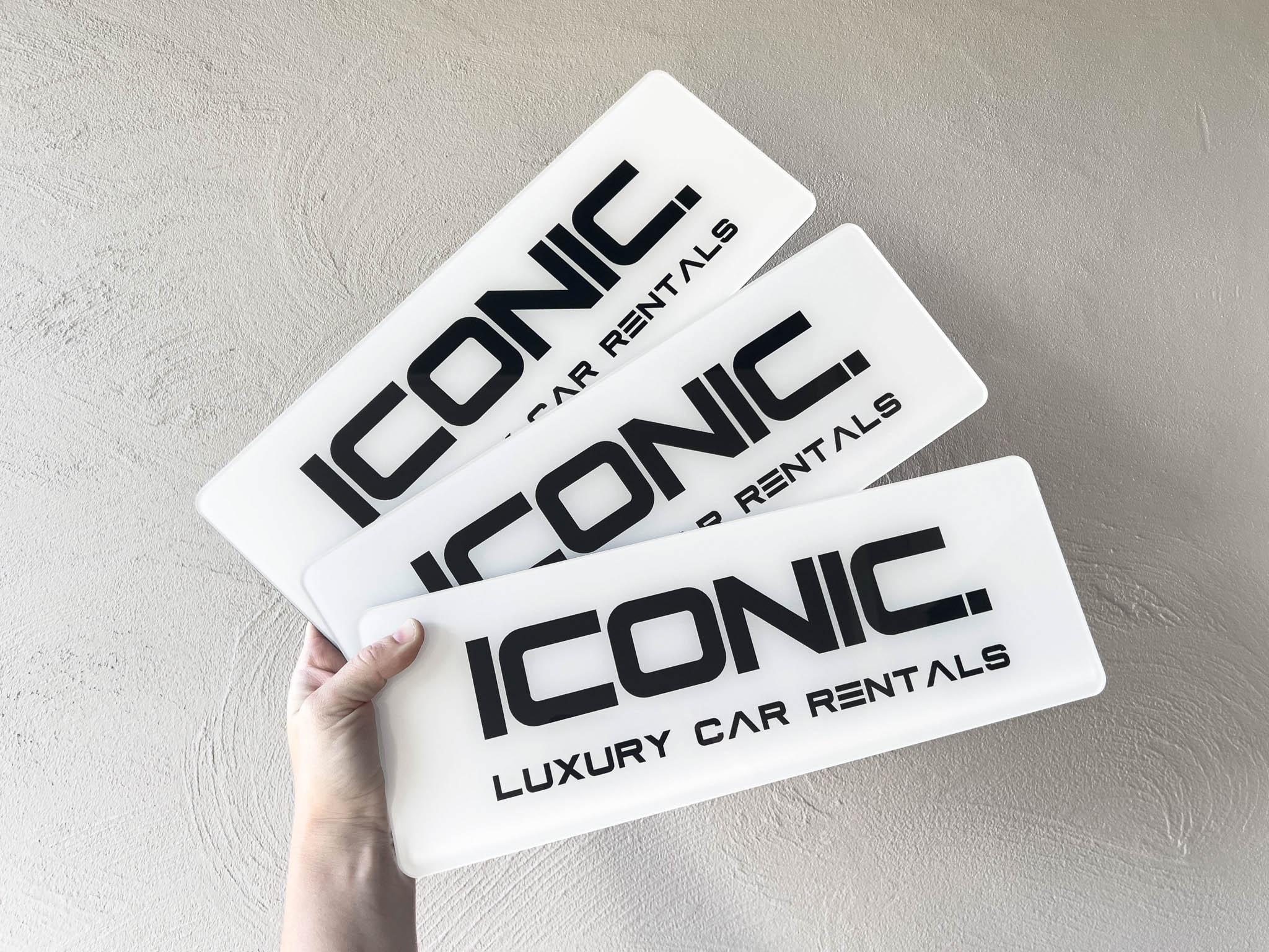 Three 'Iconic Luxury Car Rentals' glossy logo plates held by a hand on a light grey background
