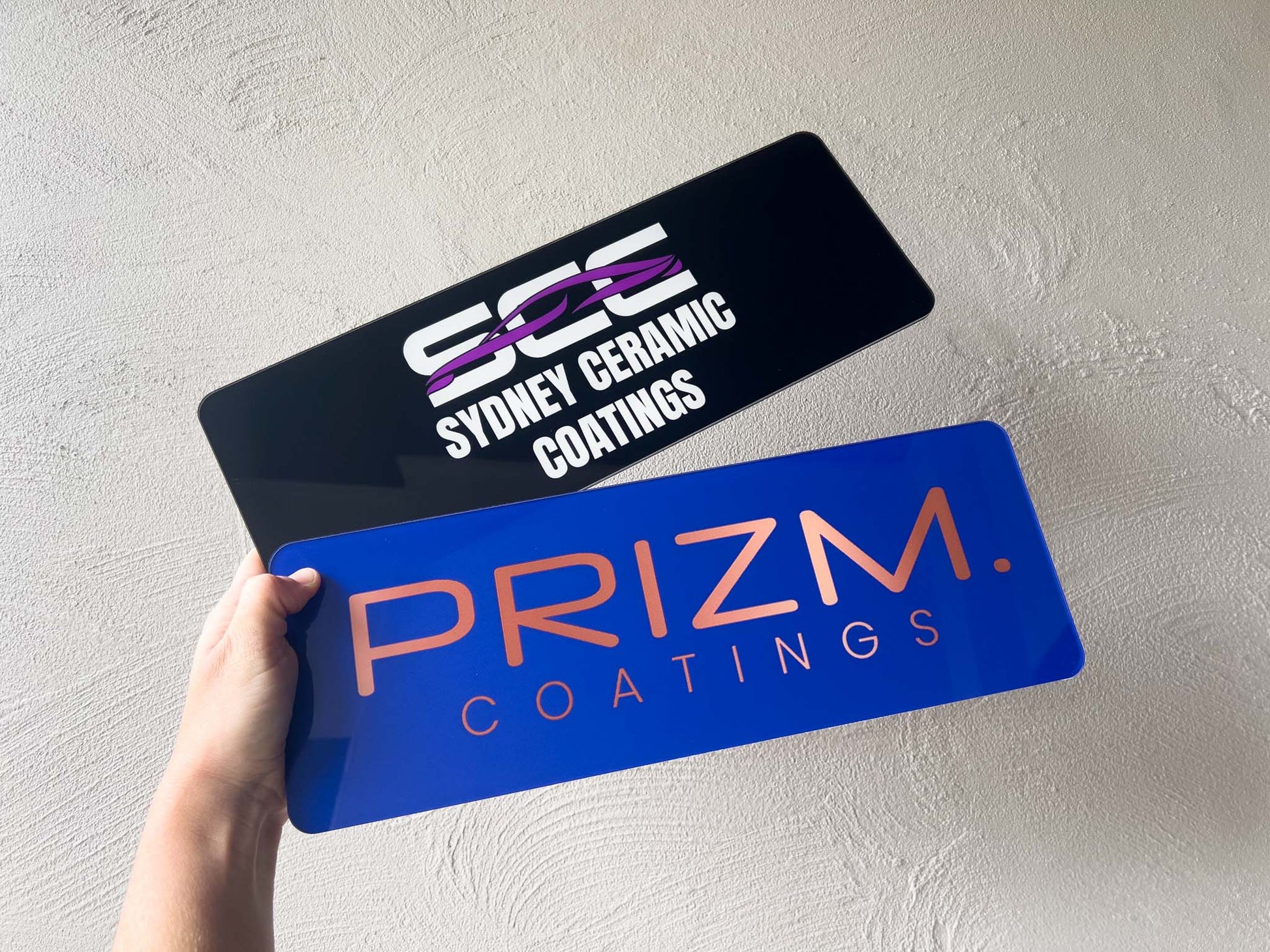 Two vehicle logo plates with 'Sydney Ceramic Coatings' and 'Prizm Coatings' logos held against a light background.