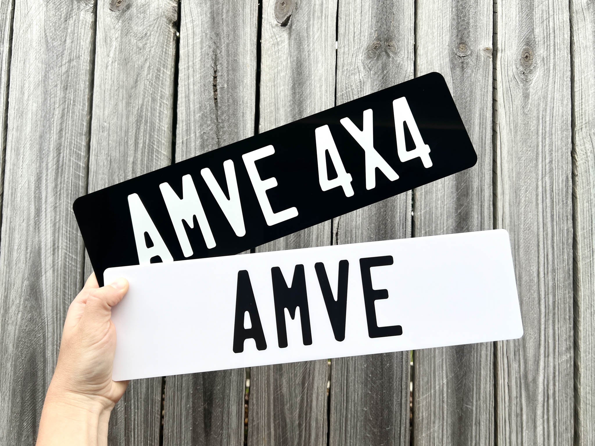 Two AMVE 4x4 acrylic license plate designs held against a wooden background