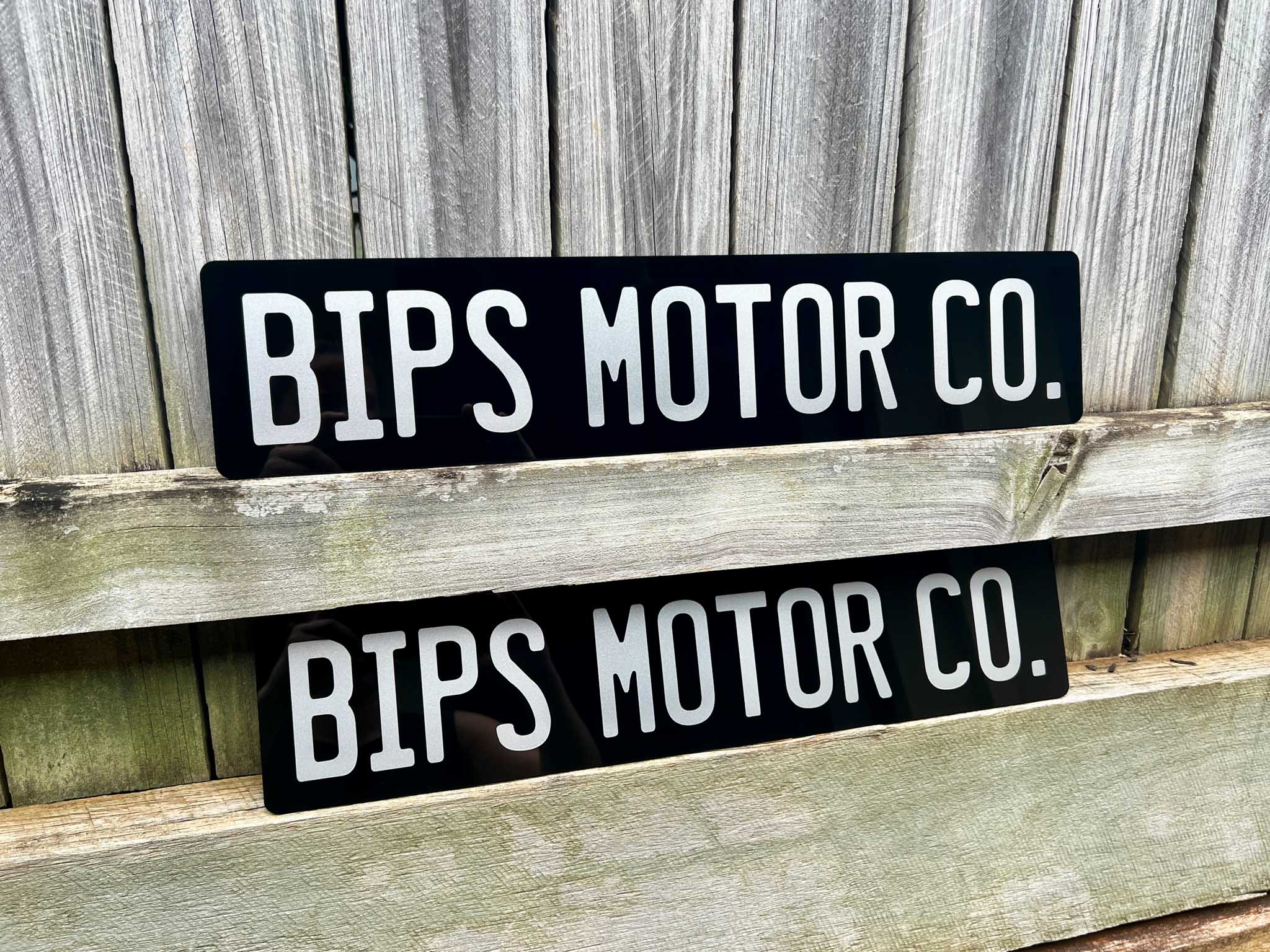 Two black and white 'BIPS MOTOR CO.' Euro acrylic signs on a wooden surface with a wooden background.