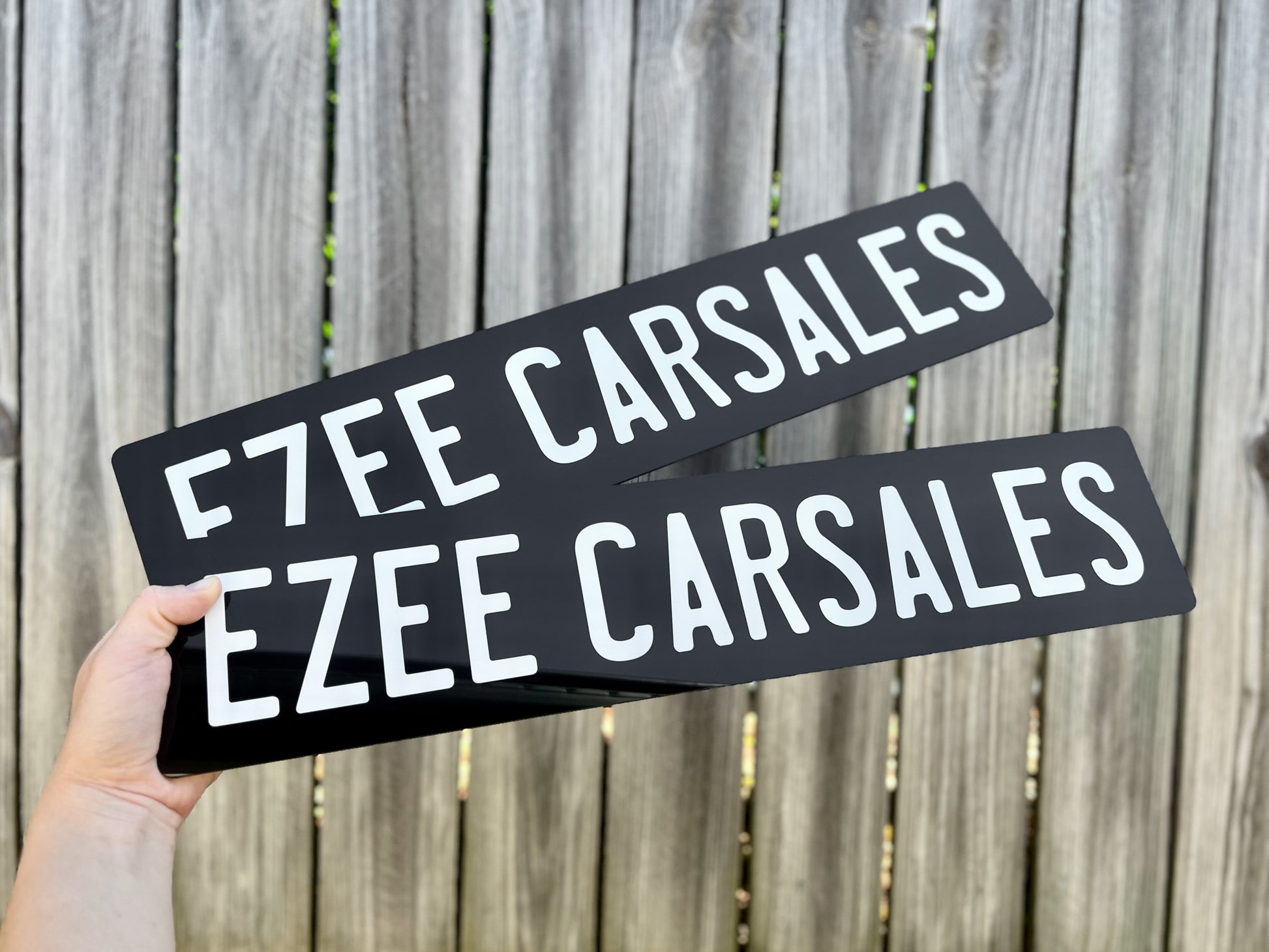 Two black Euro acrylic plates with 'EZEE CARSALES' text held against a wooden fence background.