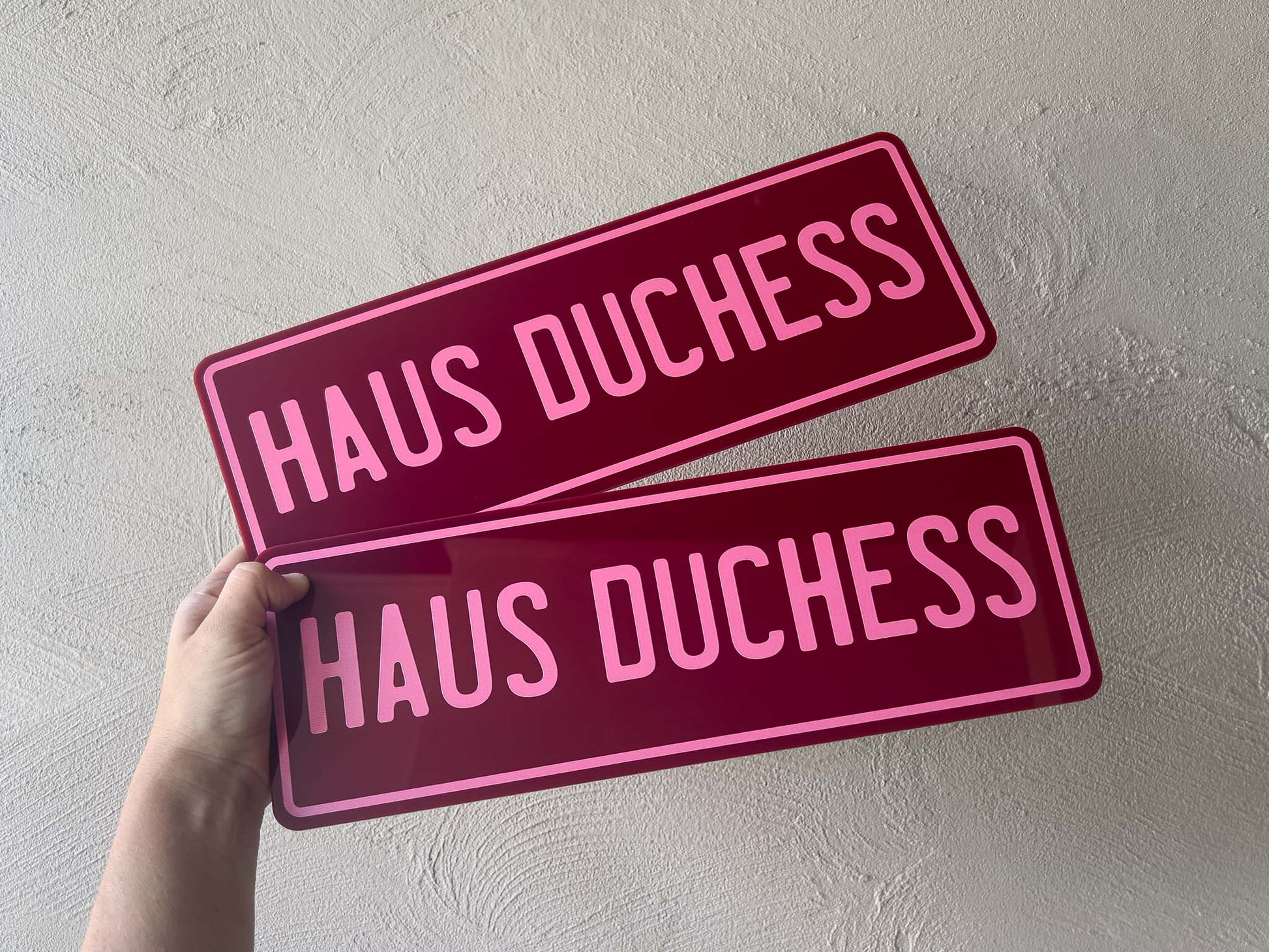 Two maroon acrylic plates with 'HAUS DUCHESS'  in pink text held against a light grey background.