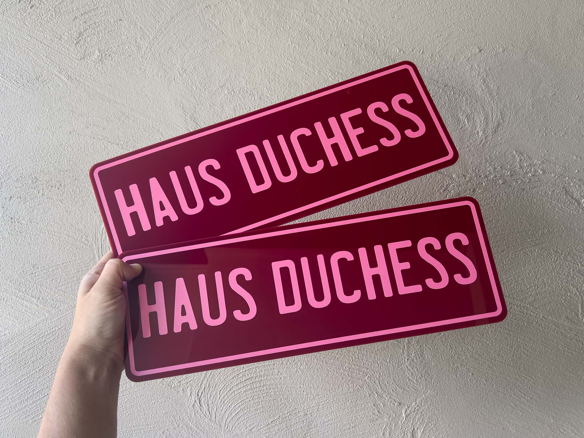 Two maroon acrylic plates with 'HAUS DUCHESS'  in pink text held against a light grey background.