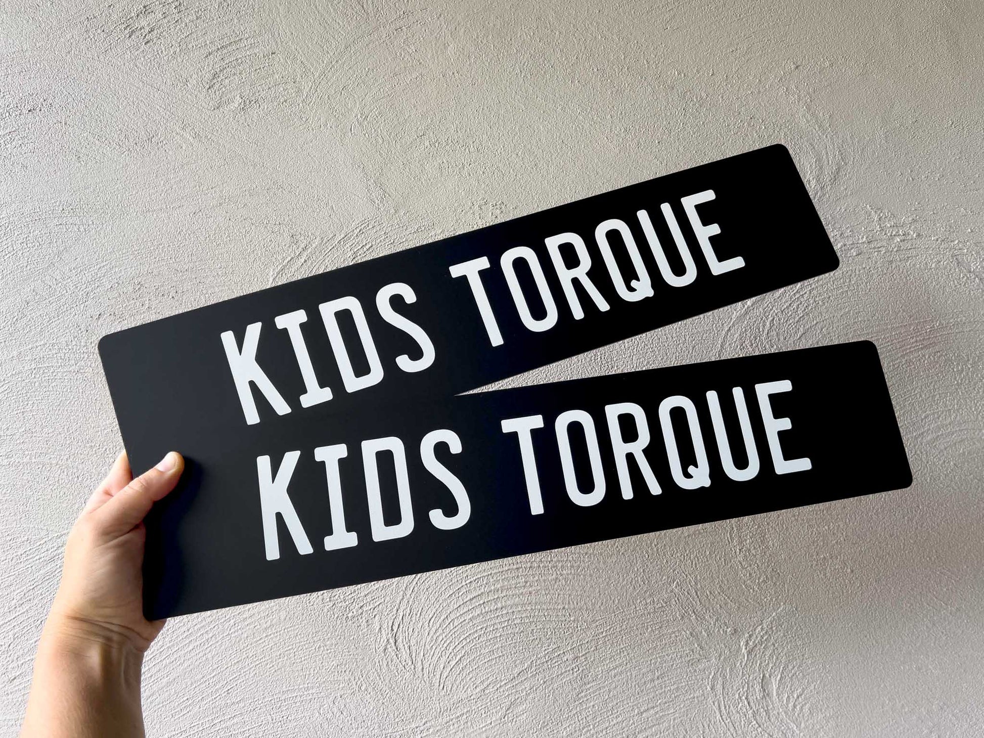 Two matte black Euro style number plates signs with 'KIDS TORQUE' text held against a light grey background.