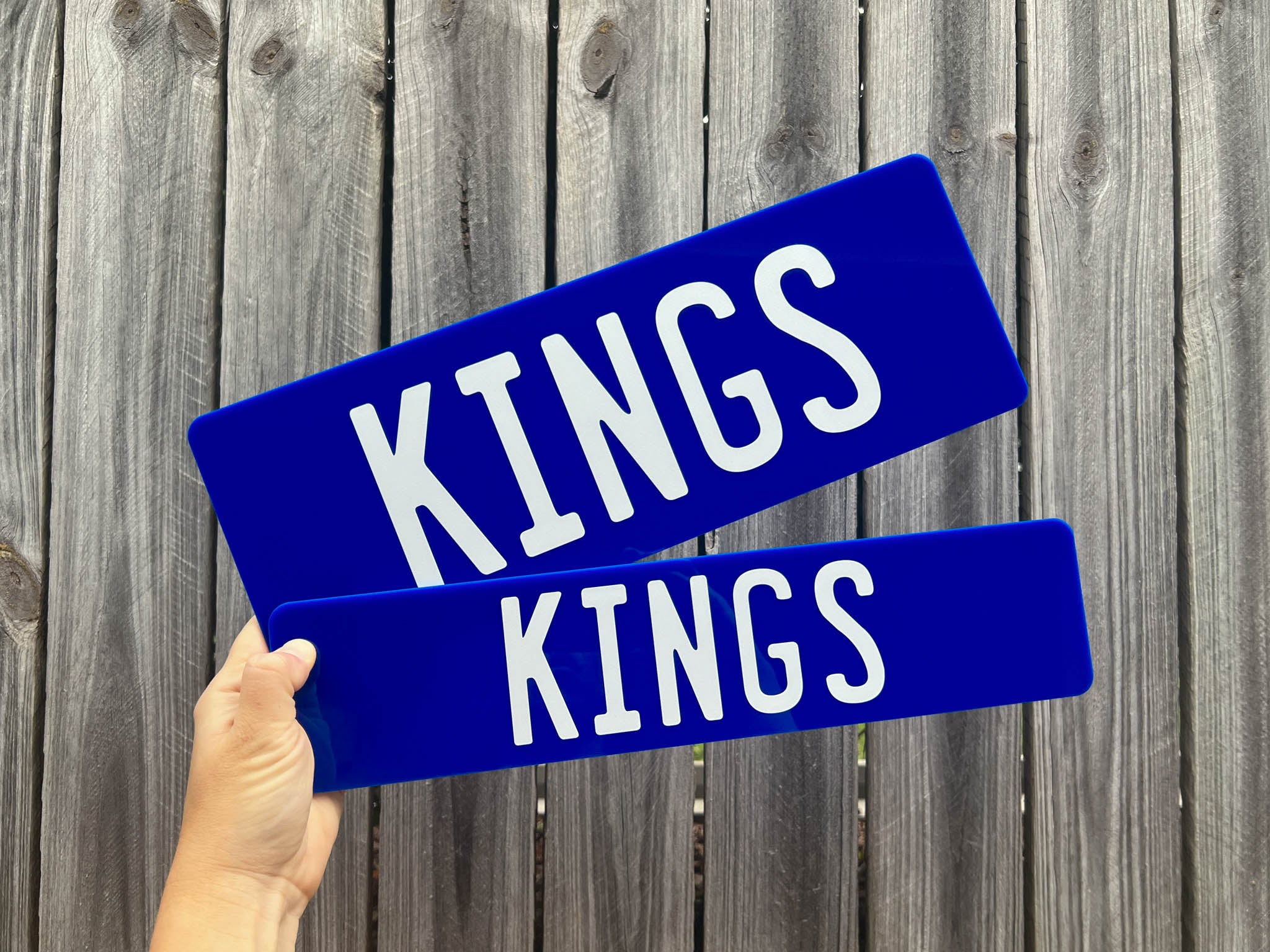 Two blue acrylic plates with 'KINGS' printed on them held against a wooden fence background.
