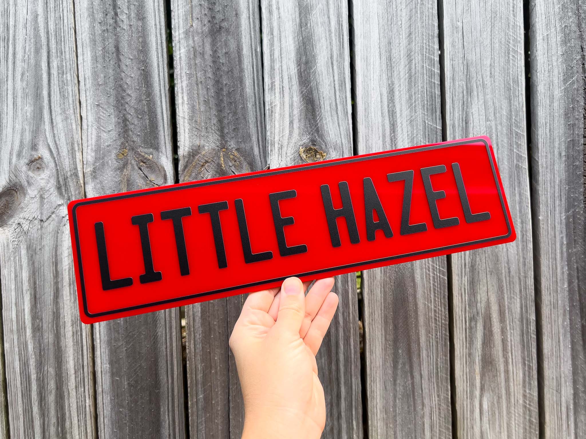 Hand holding a red slim style acrylic plate with 'LITTLE HAZEL' text against a wooden fence background