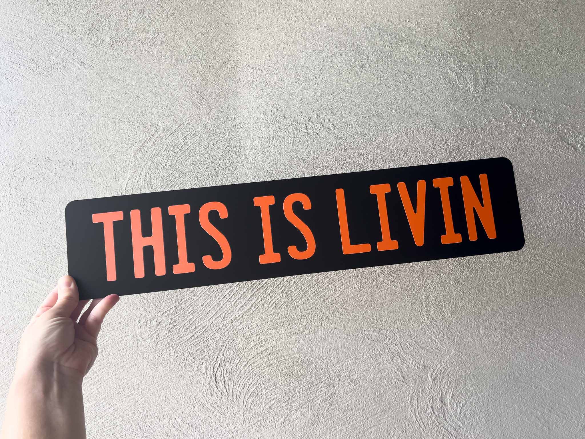 Hand holding a black Euro acrylic plate with orange text 'THIS IS LIVIN' against a light grey wall.
