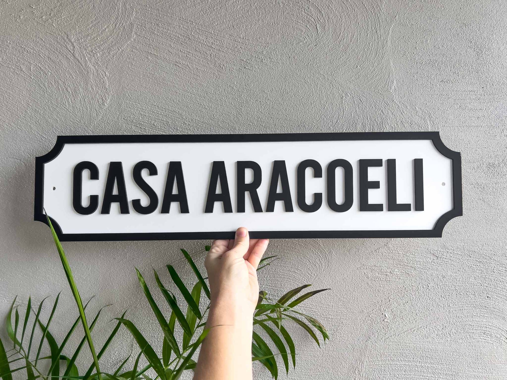 A hand holds a custom white sign with black lettering that reads 'CASA ARACOELI' against a textured grey wall, with green plant leaves visible at the bottom.