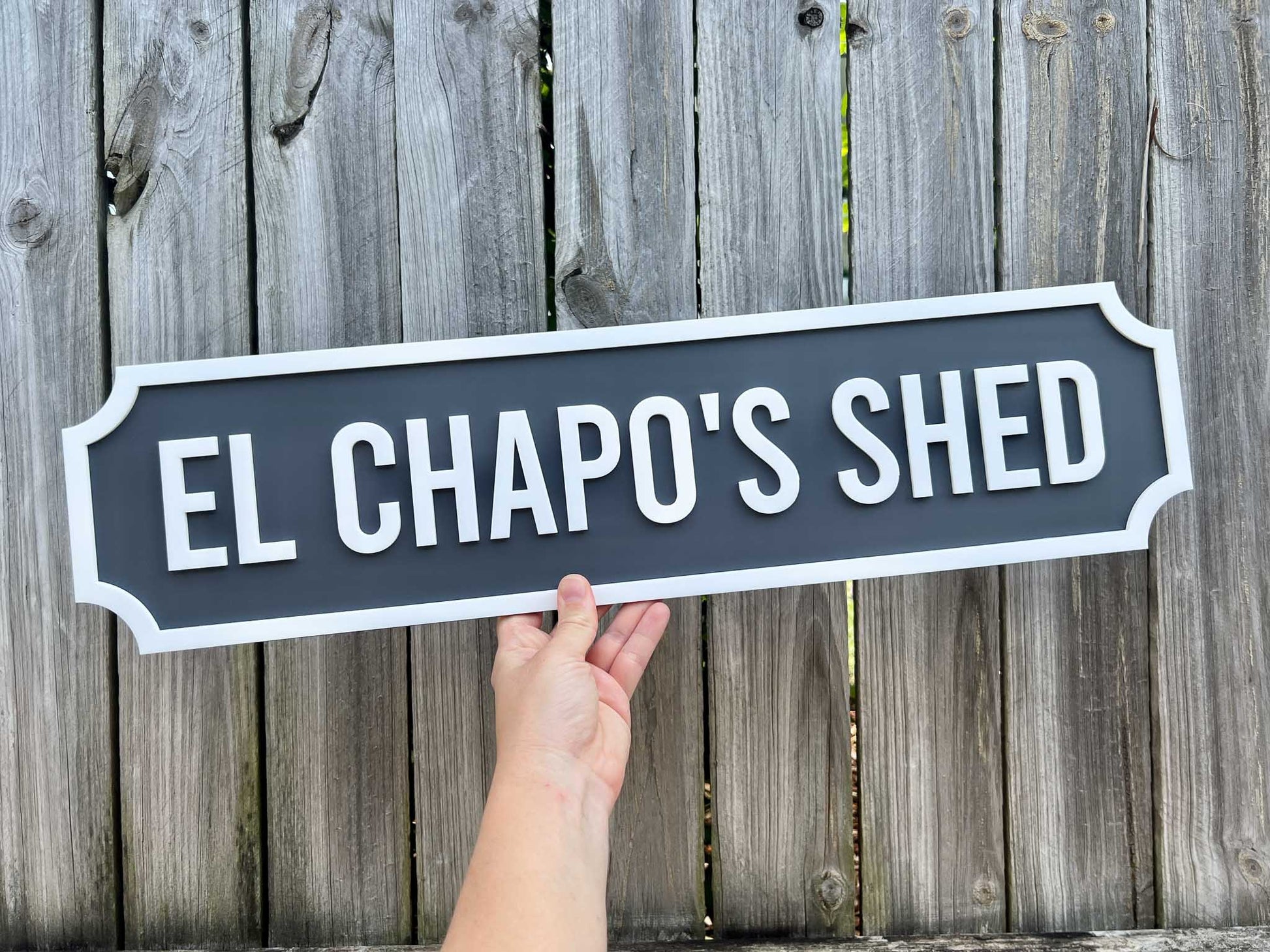 Hand holding a sign that reads 'El Chapo's Shed' against a wooden fence background