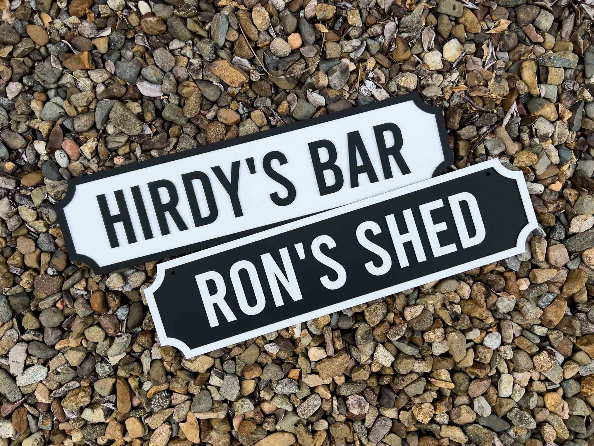 Two signs reading 'Hirdy's Bar' and 'Ron's Shed' on a bed or pebbles.