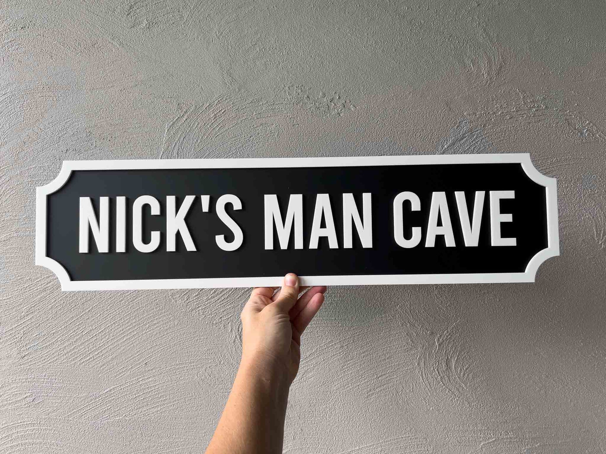 Hand holding a sign that reads 'Nick's Man Cave' against a plain background