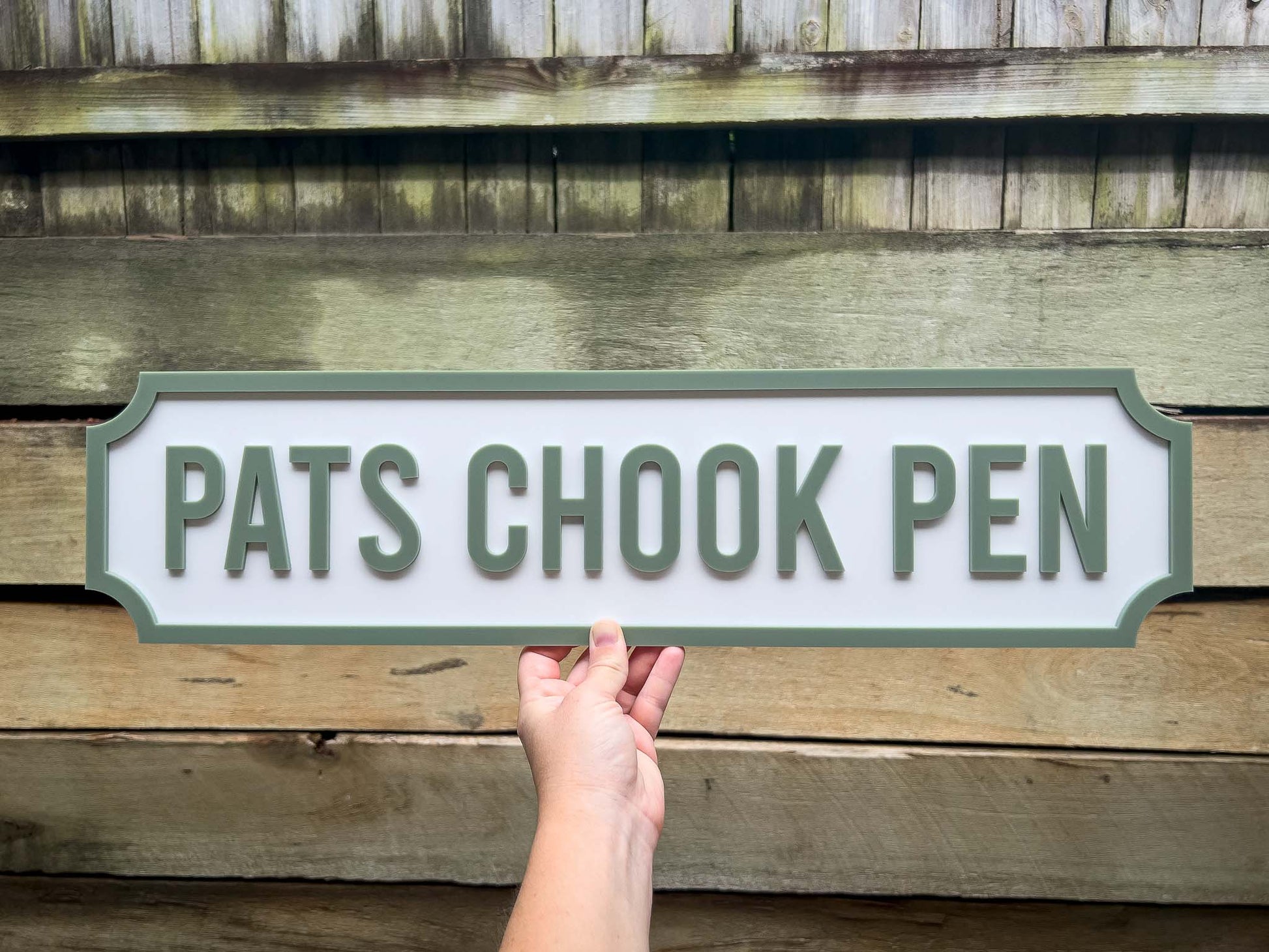 Hand holding a sign with 'PATS CHOOK PEN' text against a wooden background