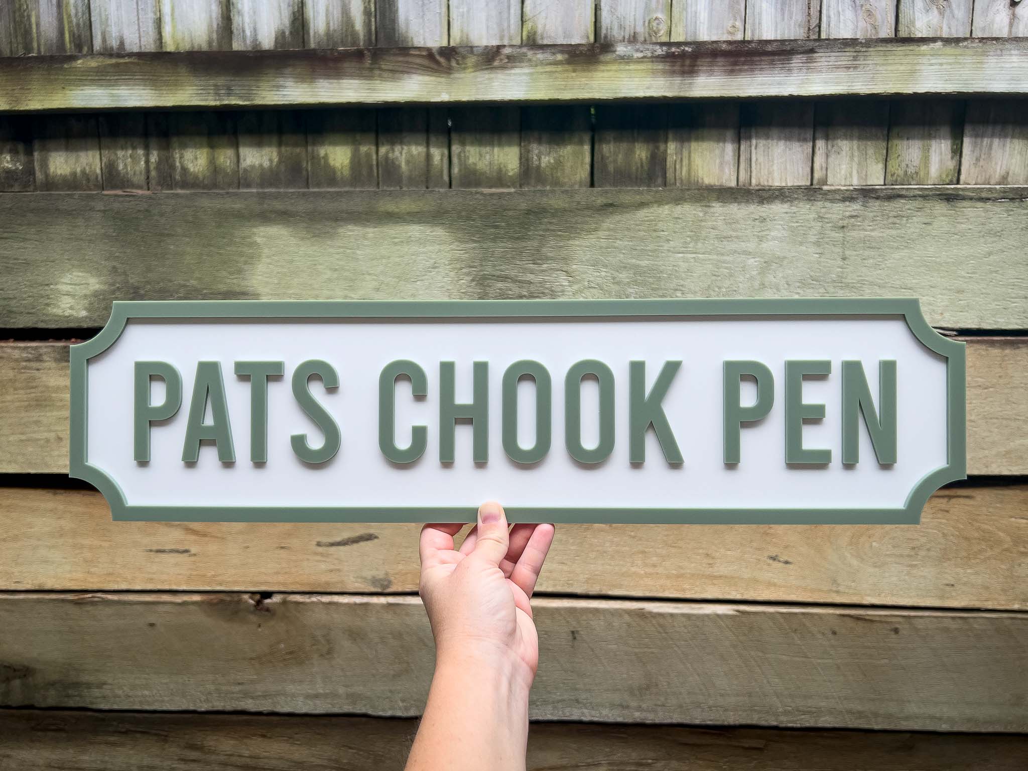 Hand holding a sign with 'PATS CHOOK PEN' text against a wooden background