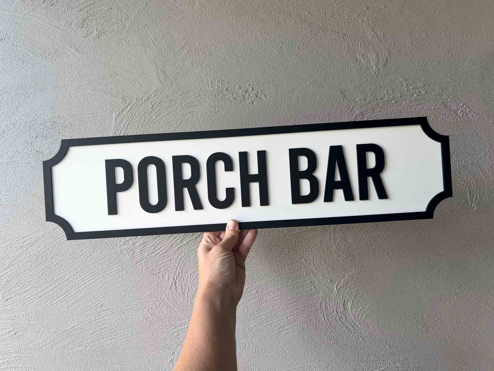 Hand holding a 'Porch Bar' sign against a plain wall