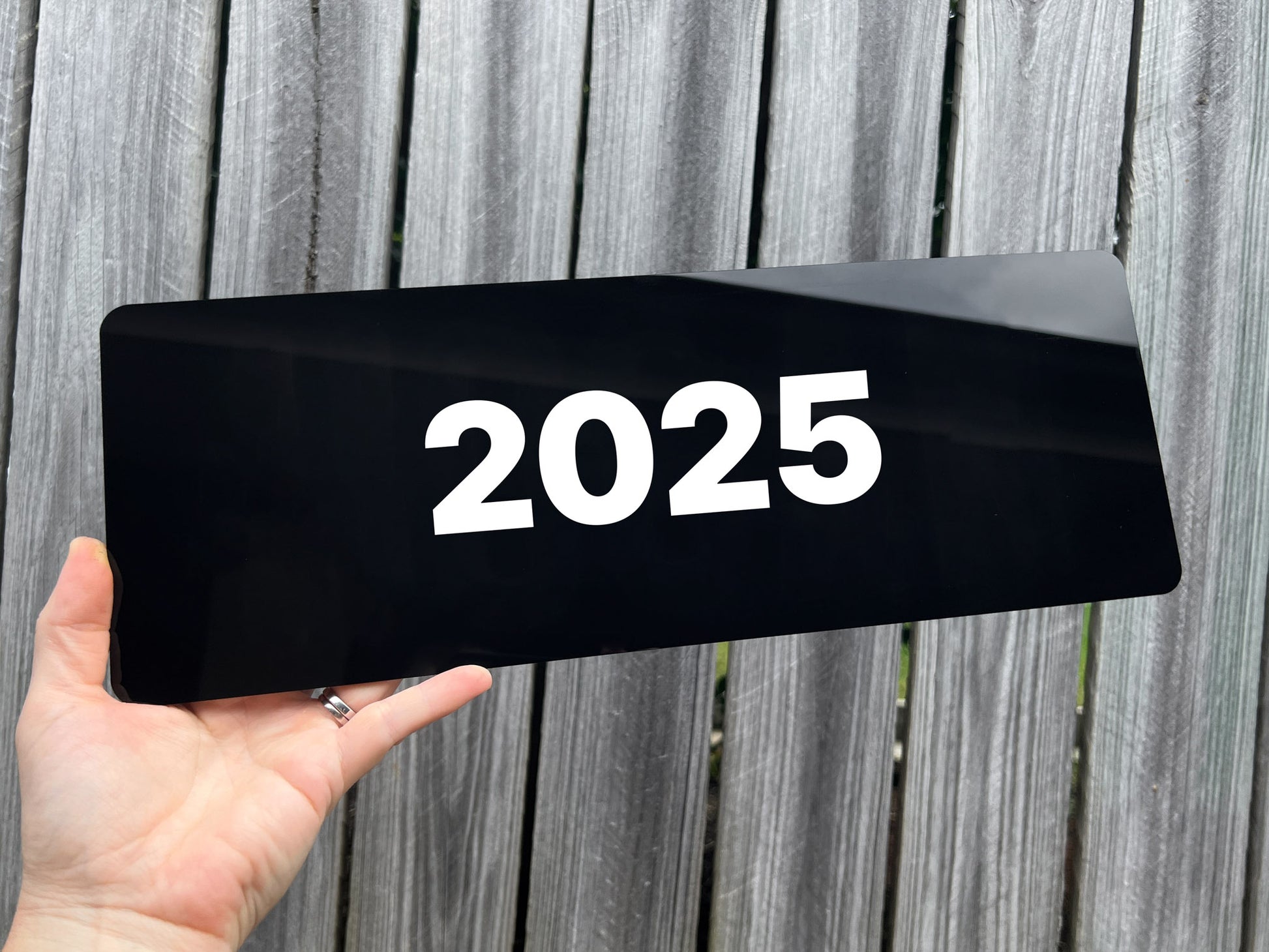 Hand holding a black gloss acrylic plate with '2025' on a wooden background
