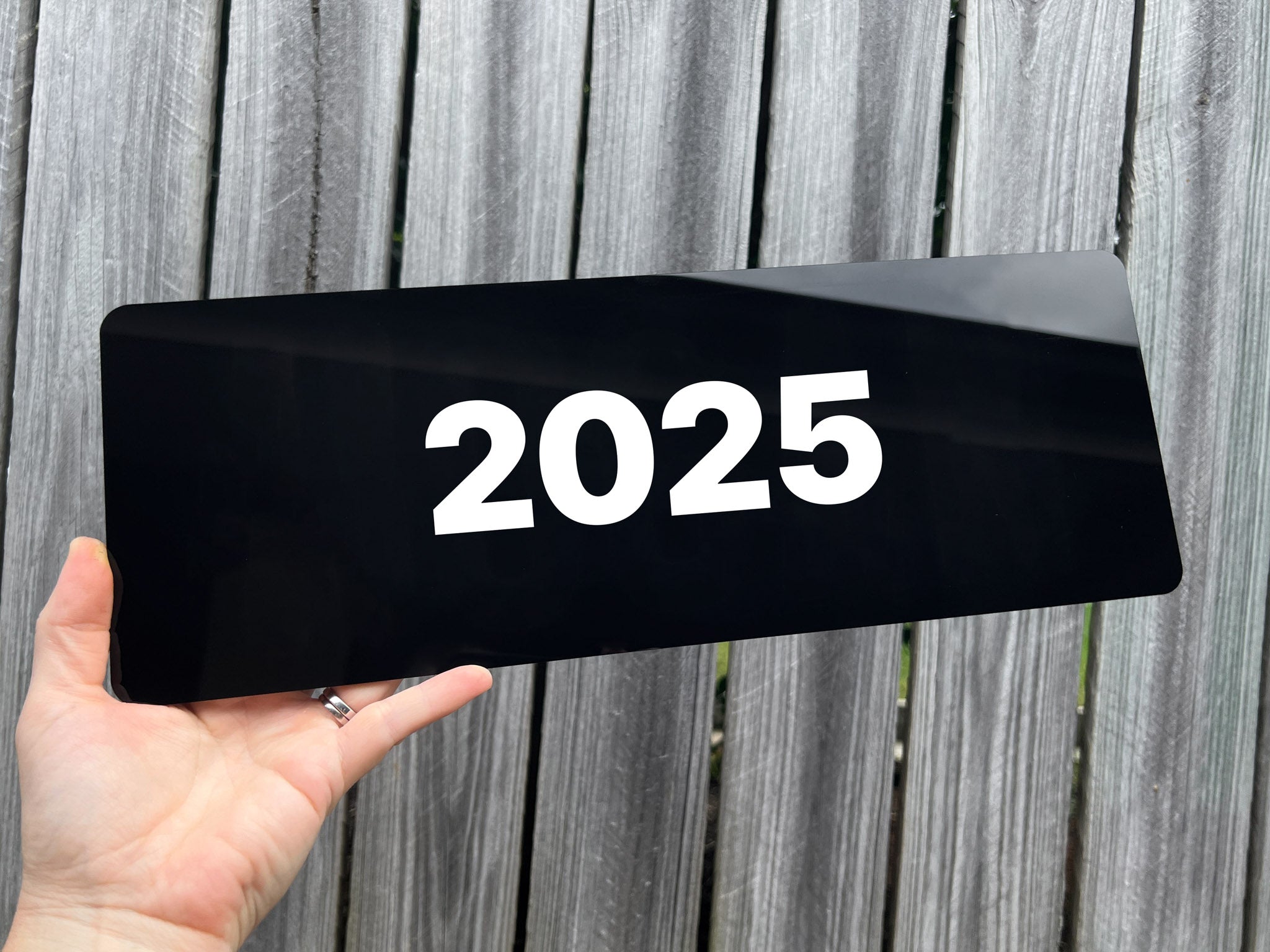 Hand holding a black gloss acrylic plate with '2025' on a wooden background