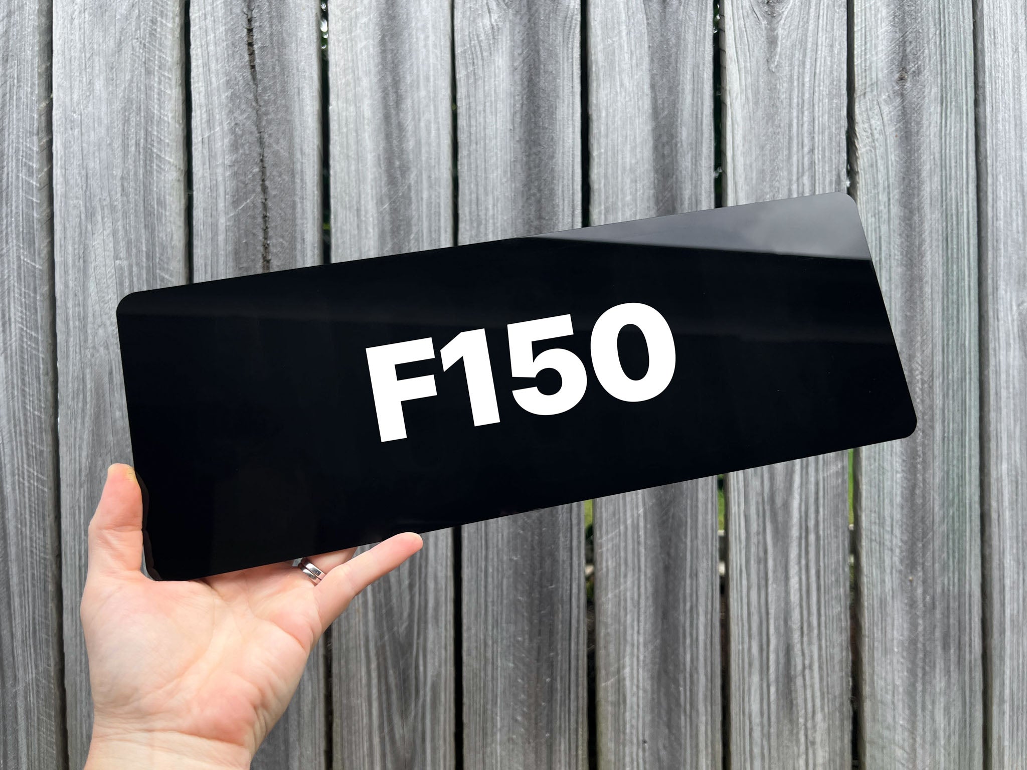Black acrylic plate with 'F150' in white text held in front of a wooden fence