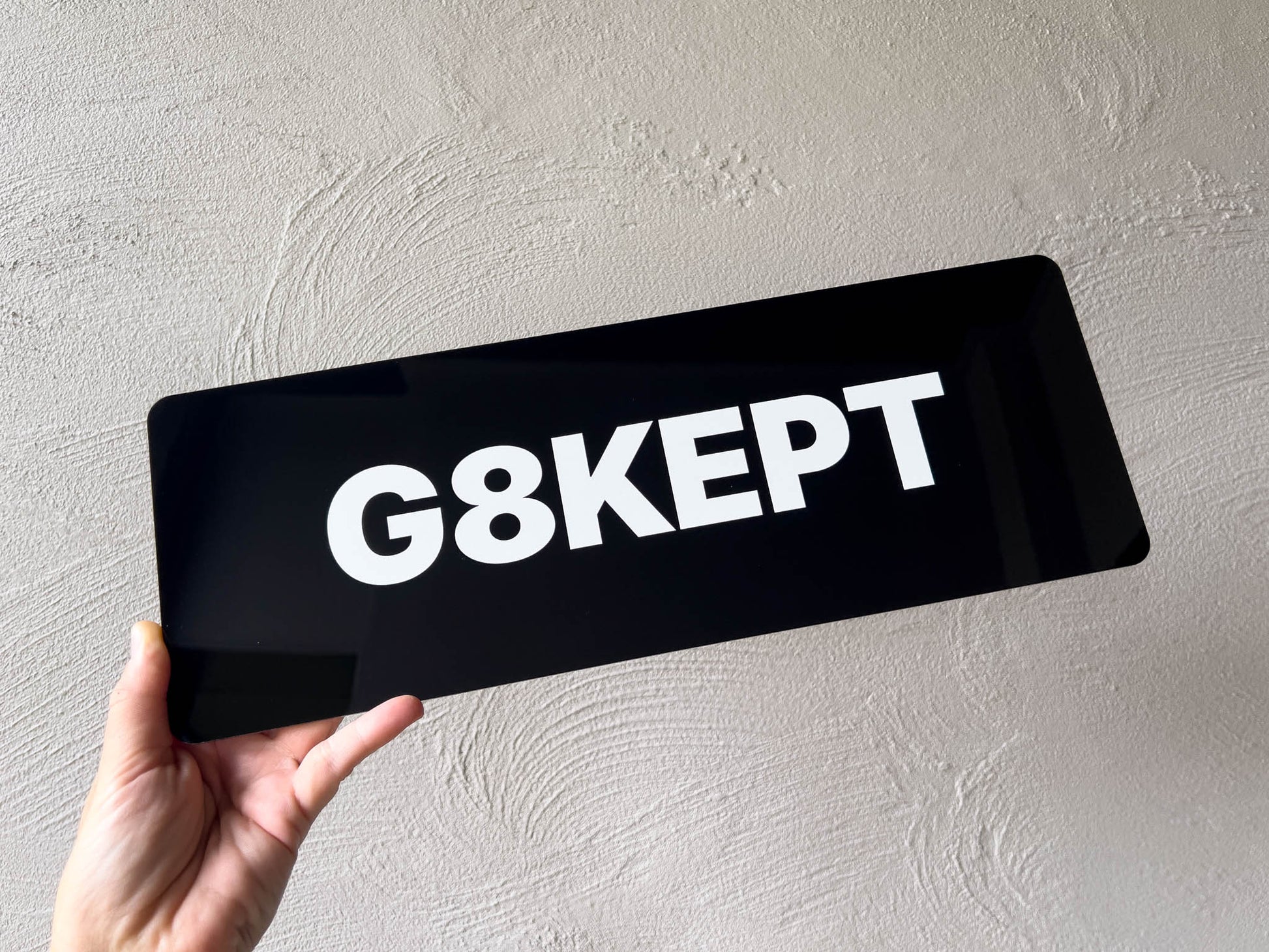 Hand holding a black acrylic plate with 'G8KEPT' text against a light grey background