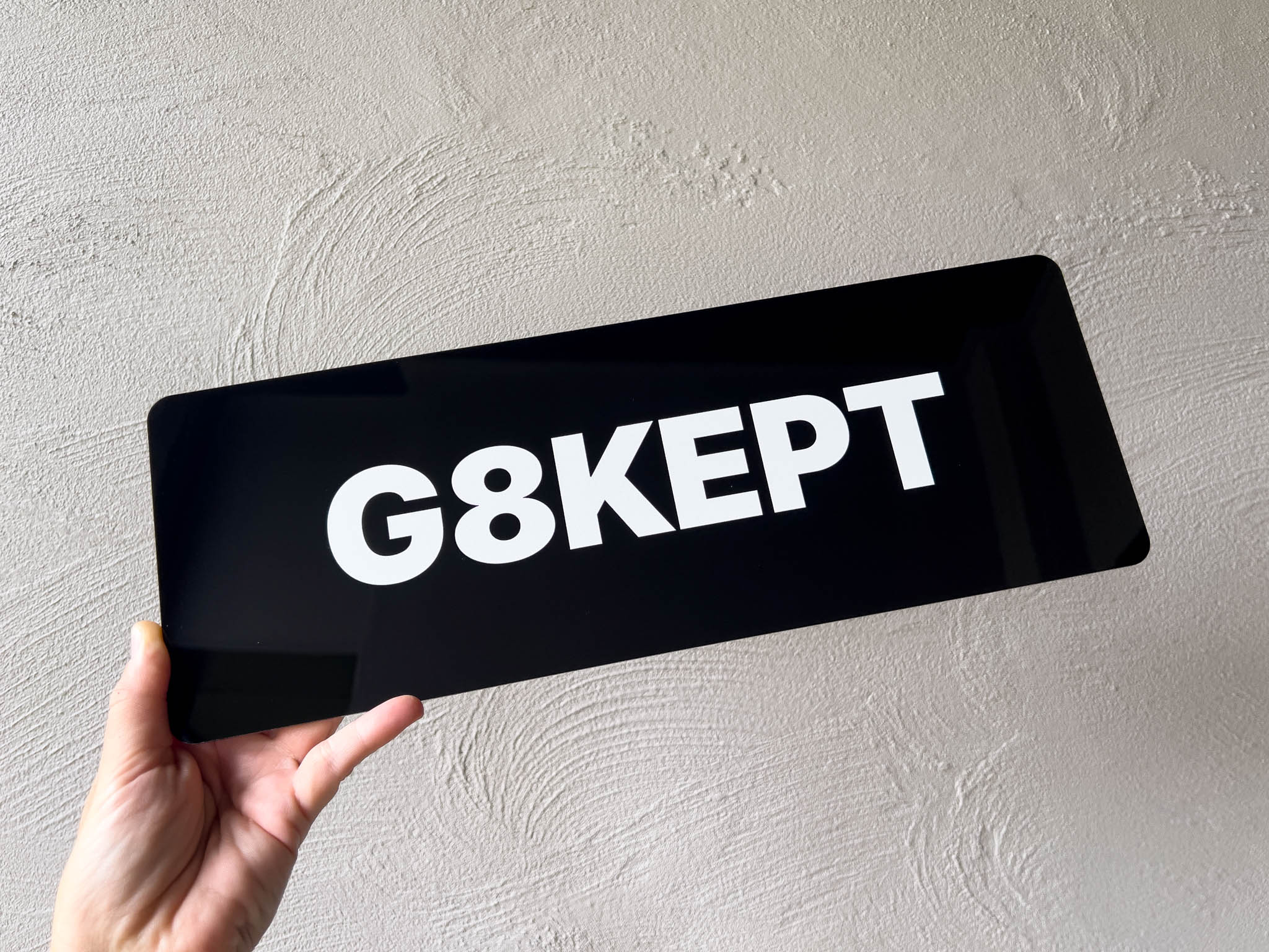 Hand holding a black acrylic plate with 'G8KEPT' text against a light grey background