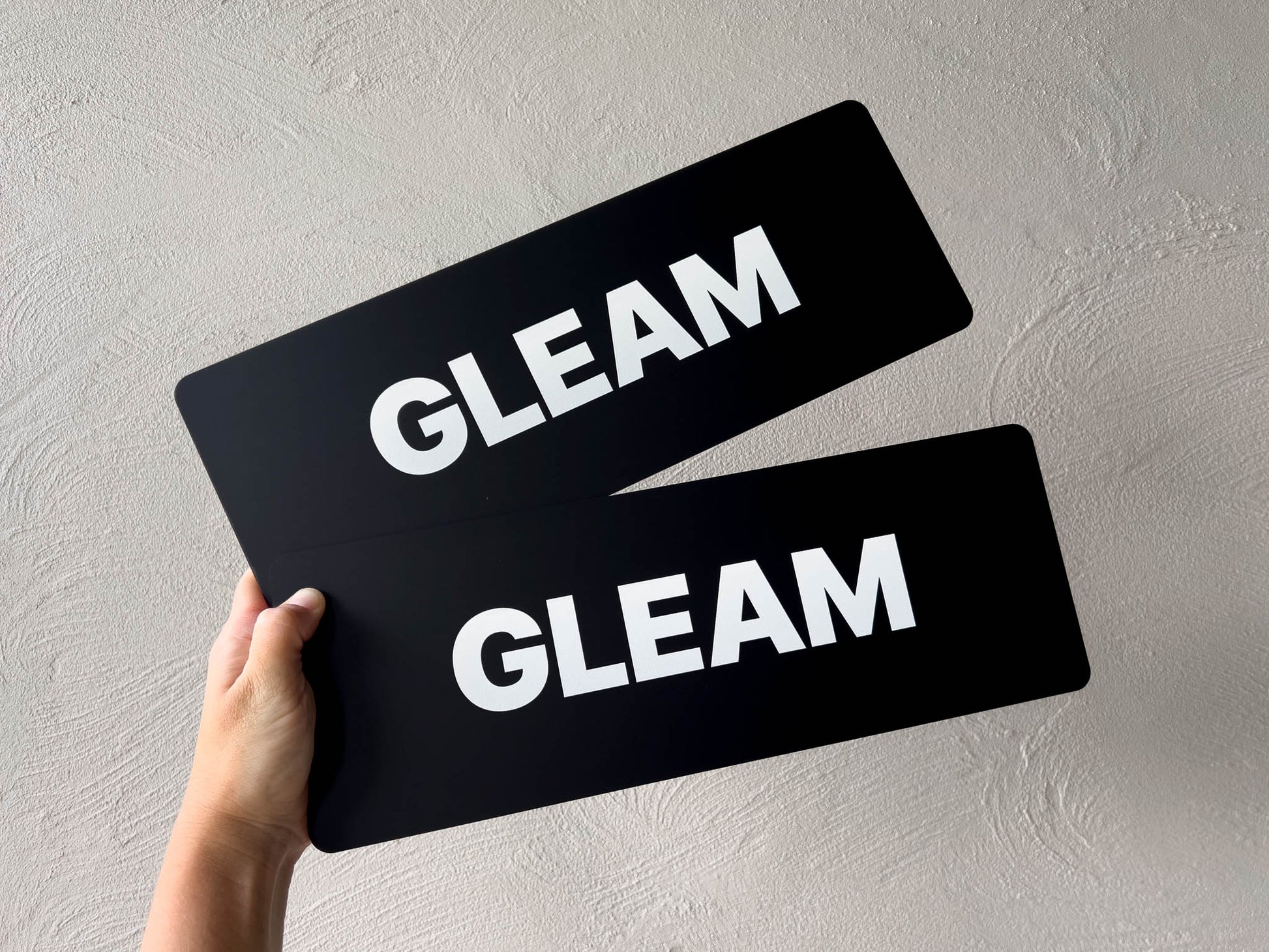 Two matte black acrylic plates with 'GLEAM' text held against a grey background