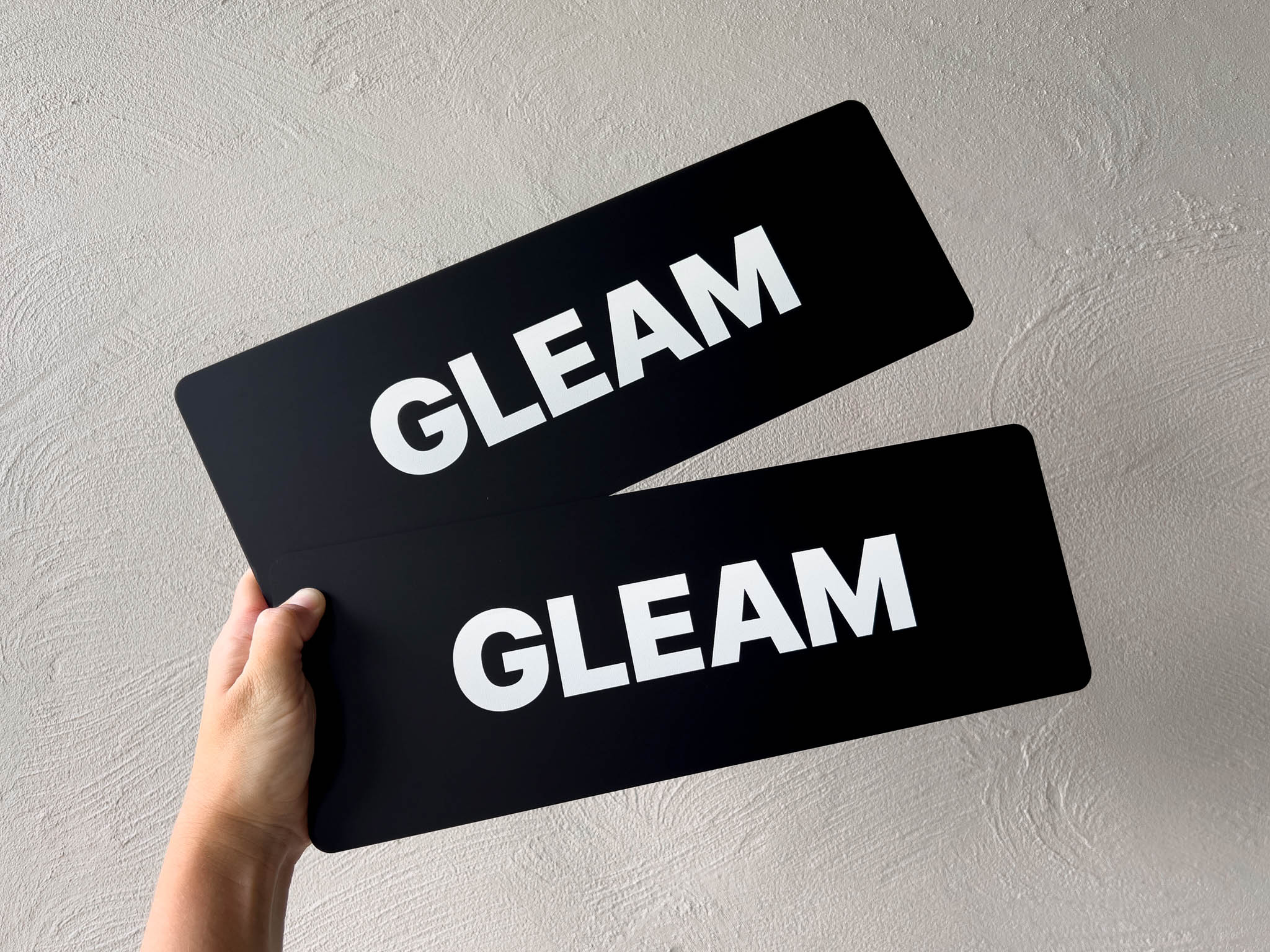 Two matte black acrylic plates with 'GLEAM' text held against a grey background