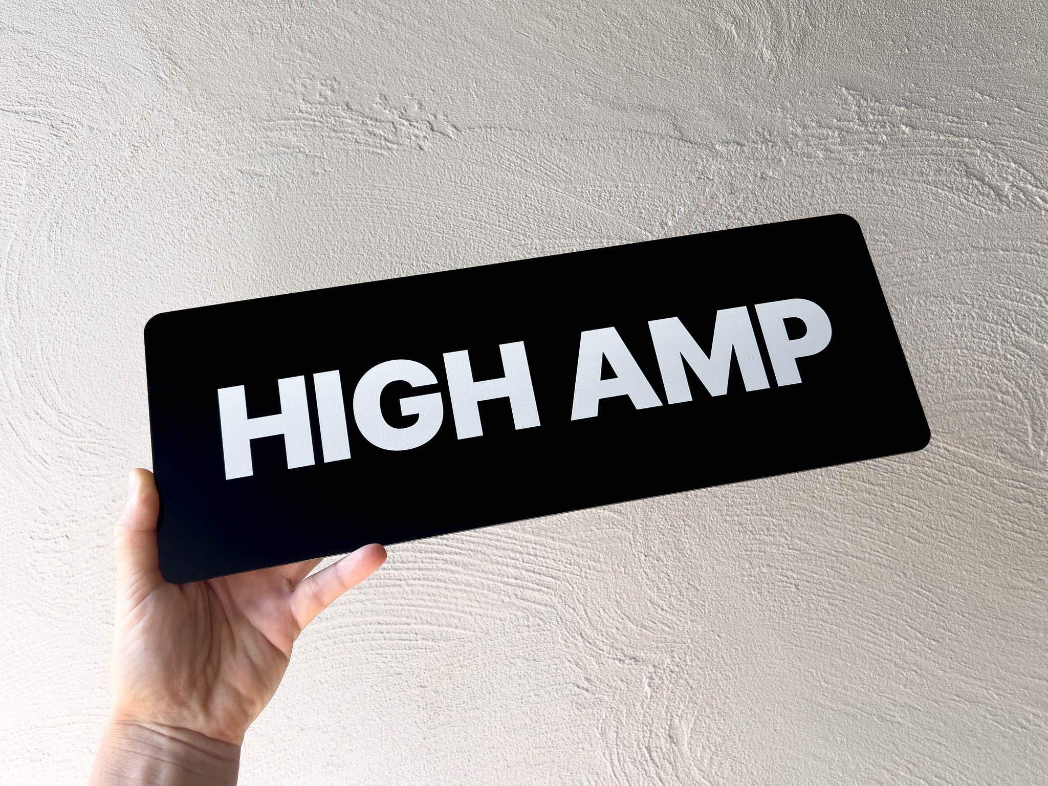 Hand holding a black sign with 'HIGH AMP' text against a light grey background