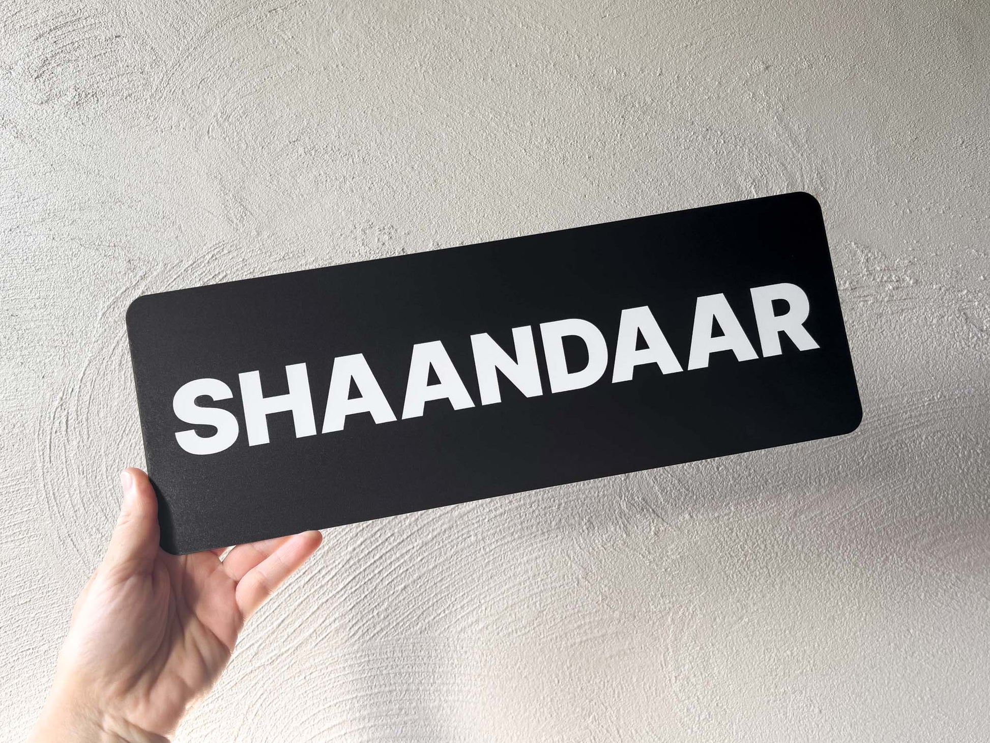 Hand holding a black sign with 'SHAANDAAR' on a light grey background