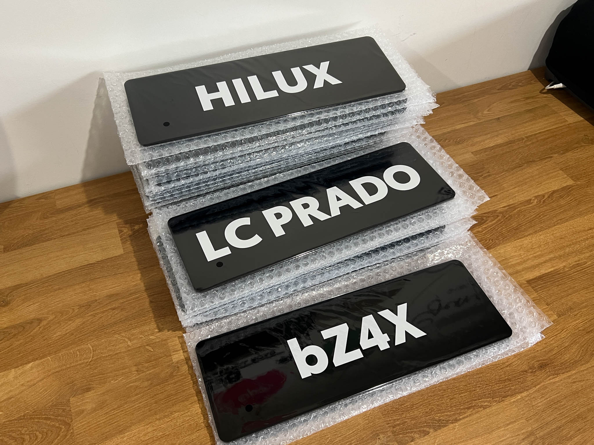 Three black glossy acrylic plates with 'HILUX', 'LC PRADO', and 'bZ4X' text on a wooden surface.