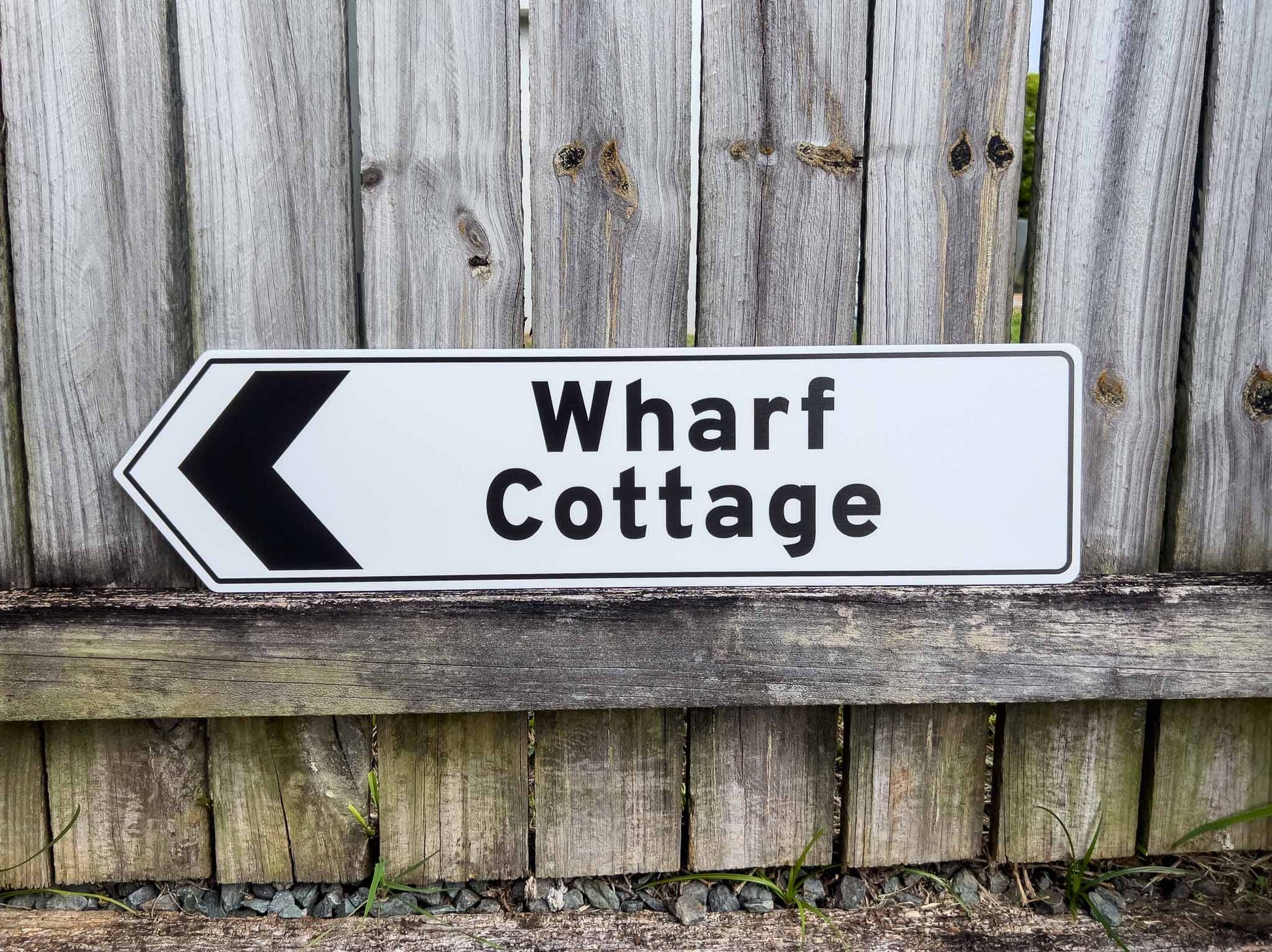 Directional sign pointing to 'Wharf Cottage' against a wooden fence background