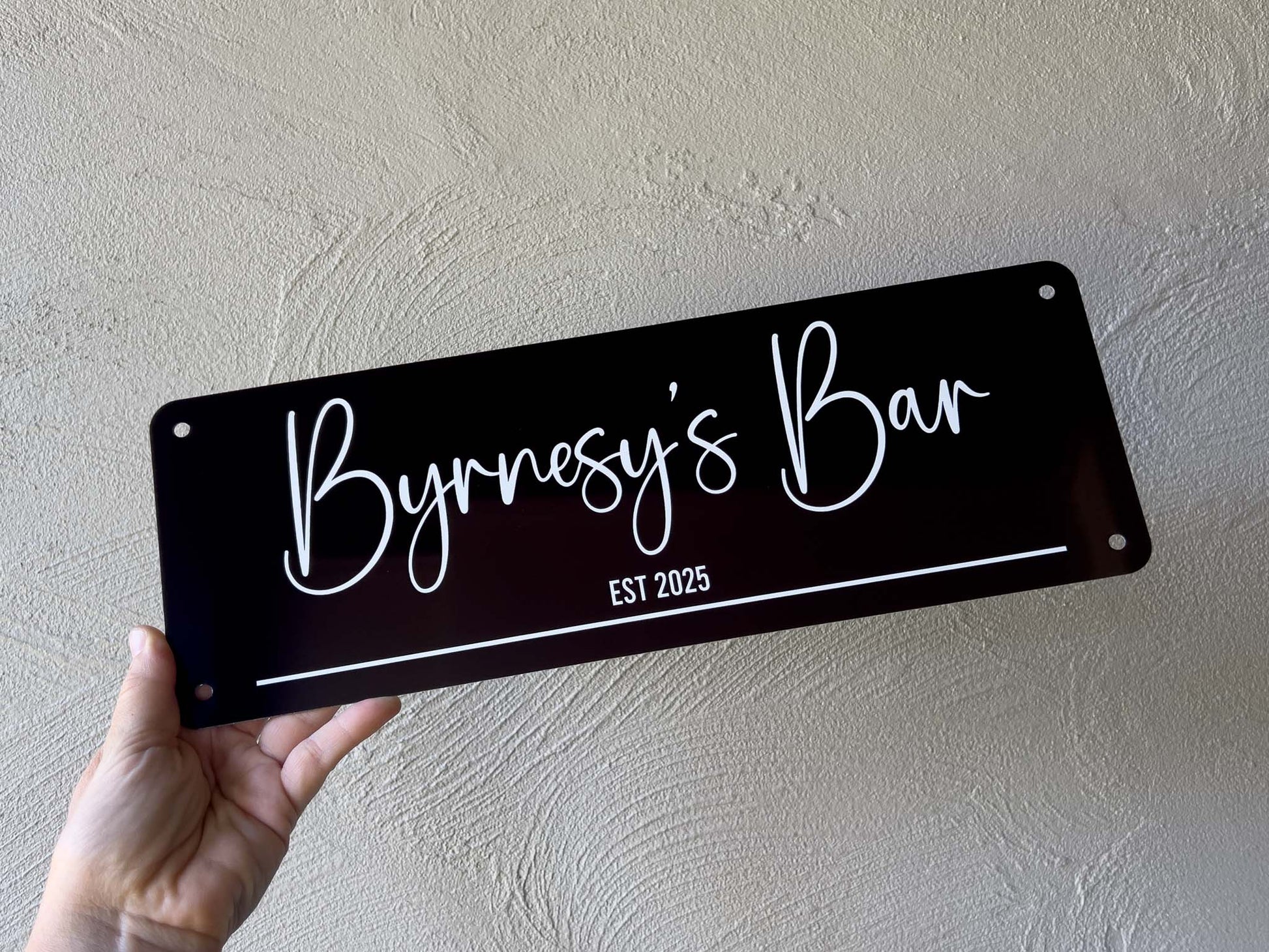 Hand holding a black sign with 'Byrnesy's Bar EST 2025' on a beige wall.
