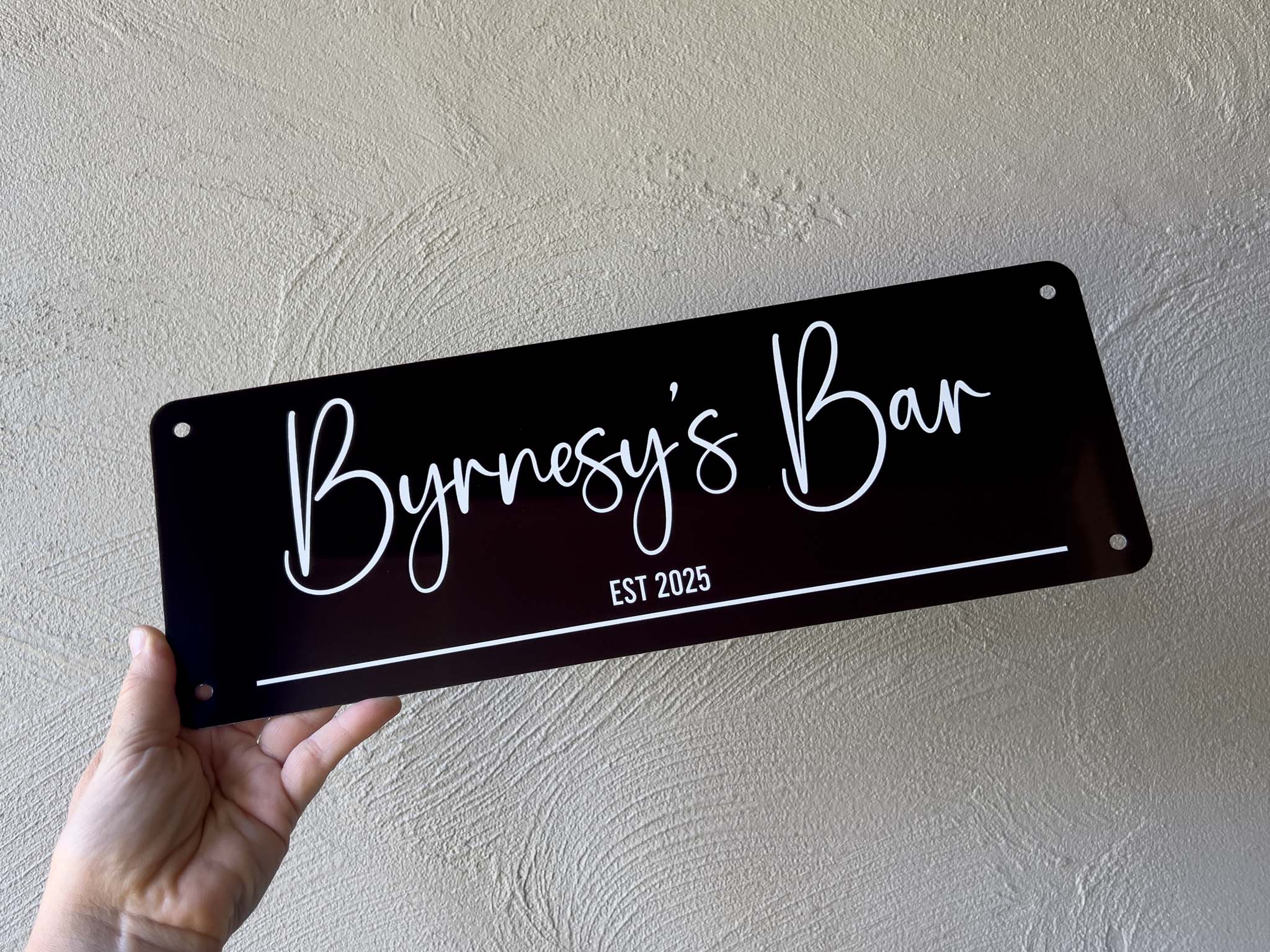Hand holding a black sign with 'Byrnesy's Bar EST 2025' on a beige wall.