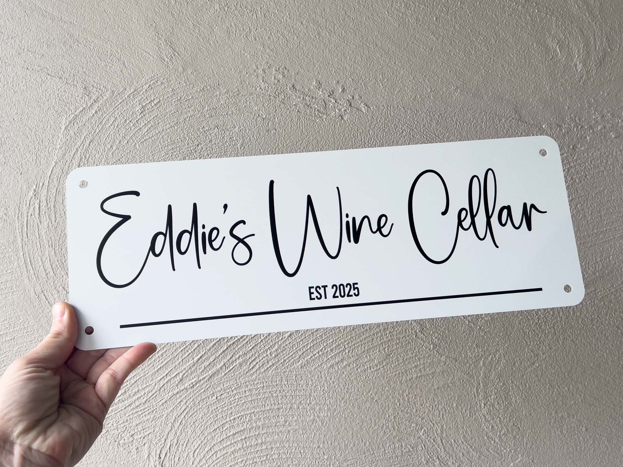 Hand holding a sign with 'Eddie's Wine Cellar EST 2025' text against a light grey wall.