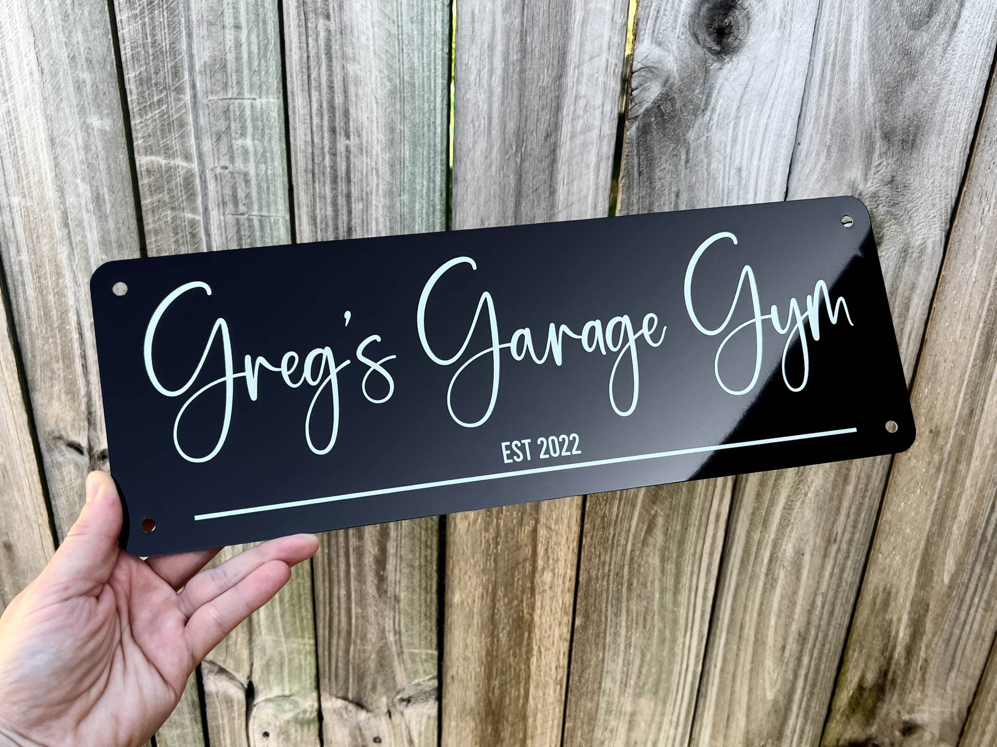 Hand holding a custom printed sign with 'Greg's Garage Gym EST 2022' text against a wooden background