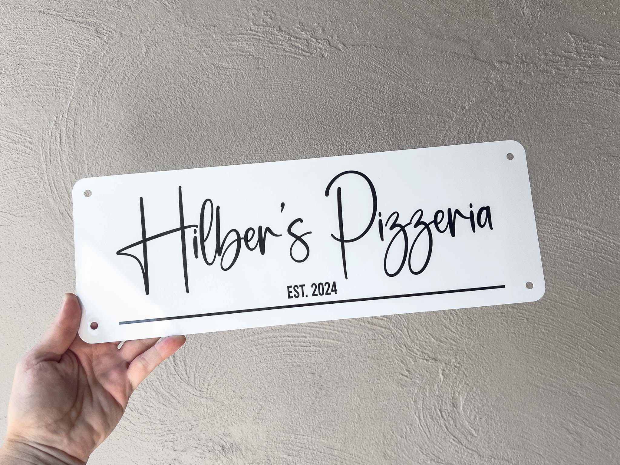 Hand holding a white metal sign with 'Hilber's Pizzeria EST. 2024' on a grey background