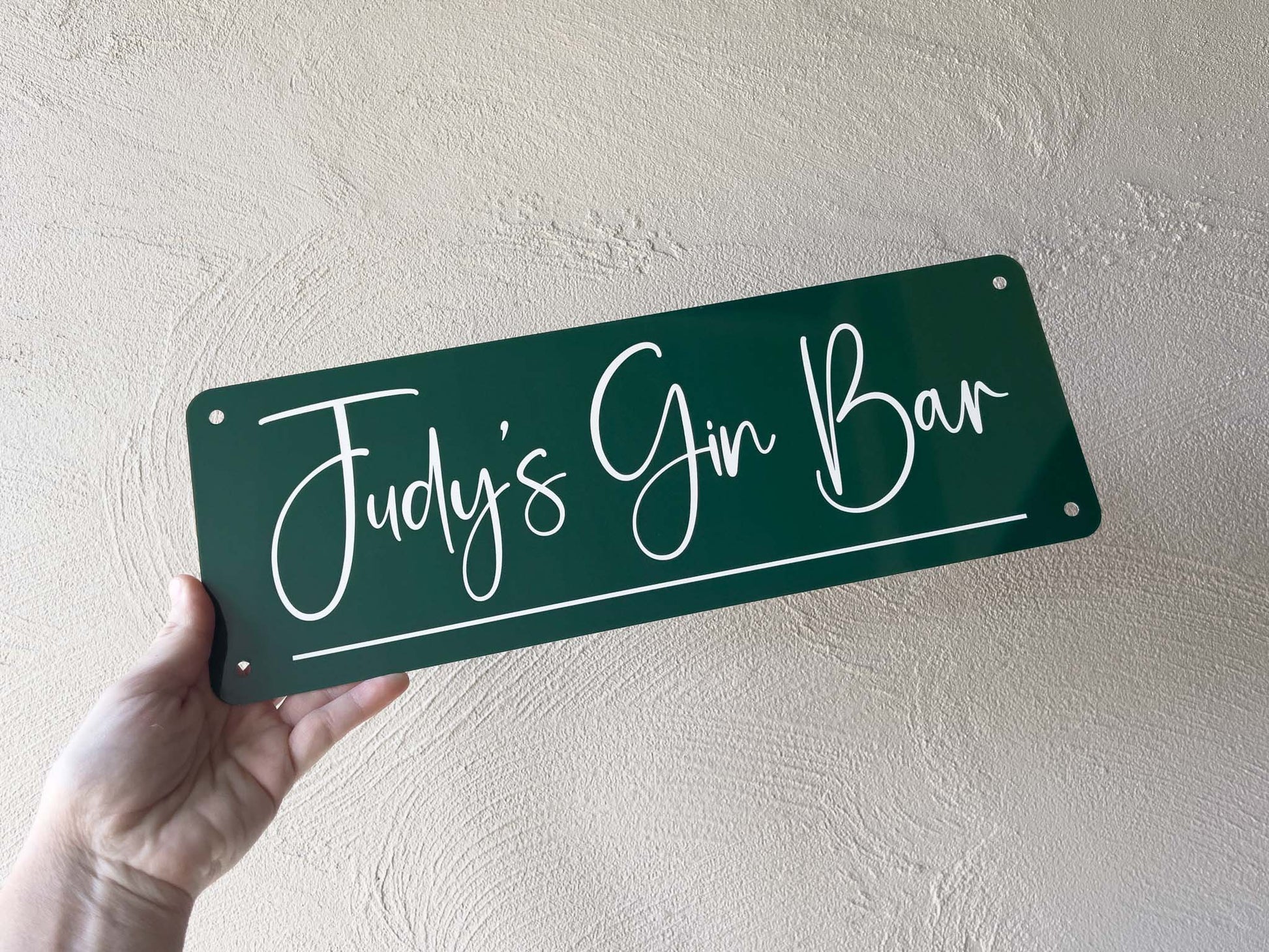 Hand holding a dark green sign with 'Judy's Gin Bar' text against a light beige wall.