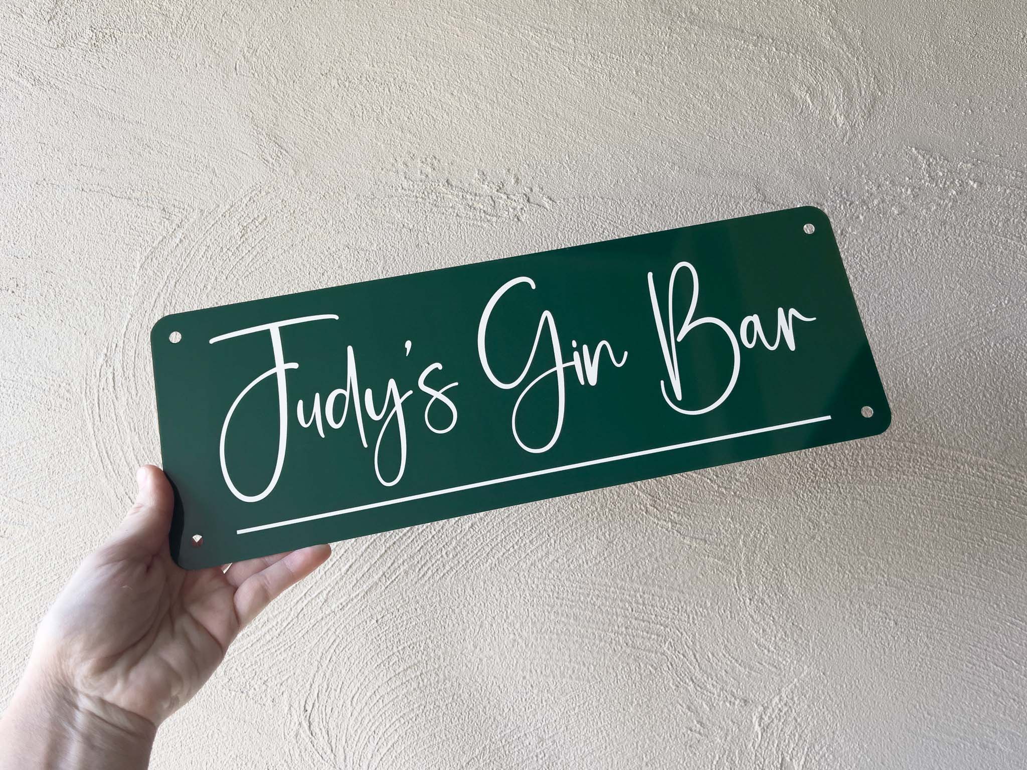 Hand holding a dark green sign with 'Judy's Gin Bar' text against a light beige wall.
