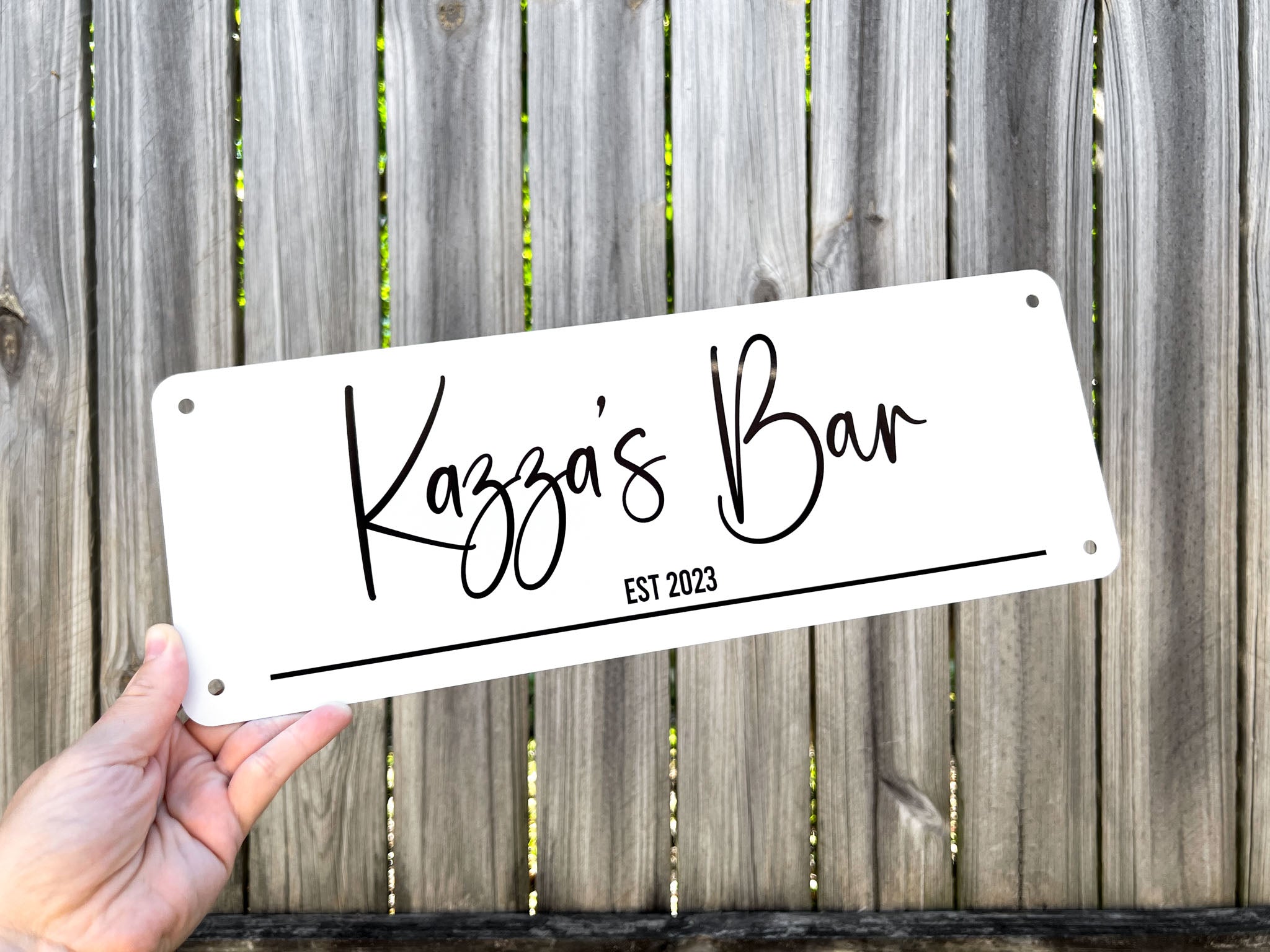 A custom printed bar style sign with 'Kazzza's Bar EST 2023' against a wooden fence background